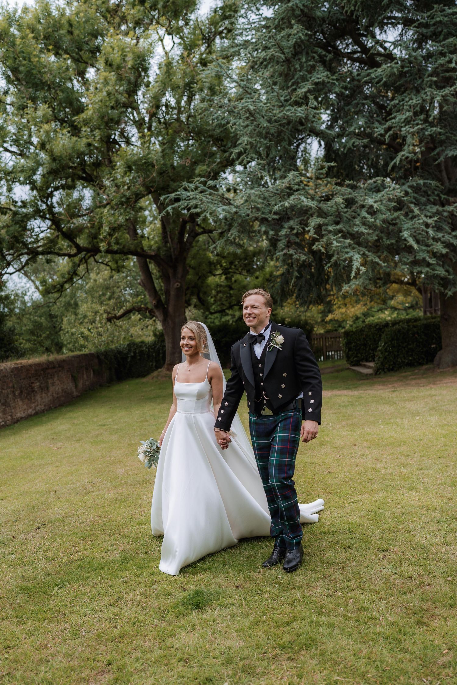 A couple walks together on a grassy hillside during their wedding celebration in traditional Scottish attire.