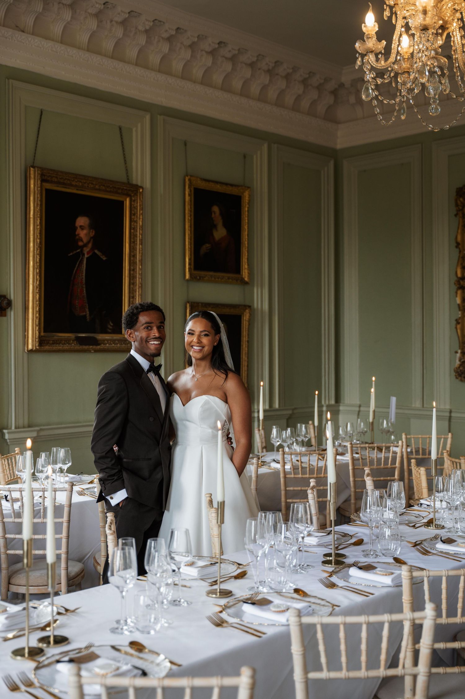Elegant wedding reception venue with crystal chandeliers and white place settings in a historic room with mint green walls.