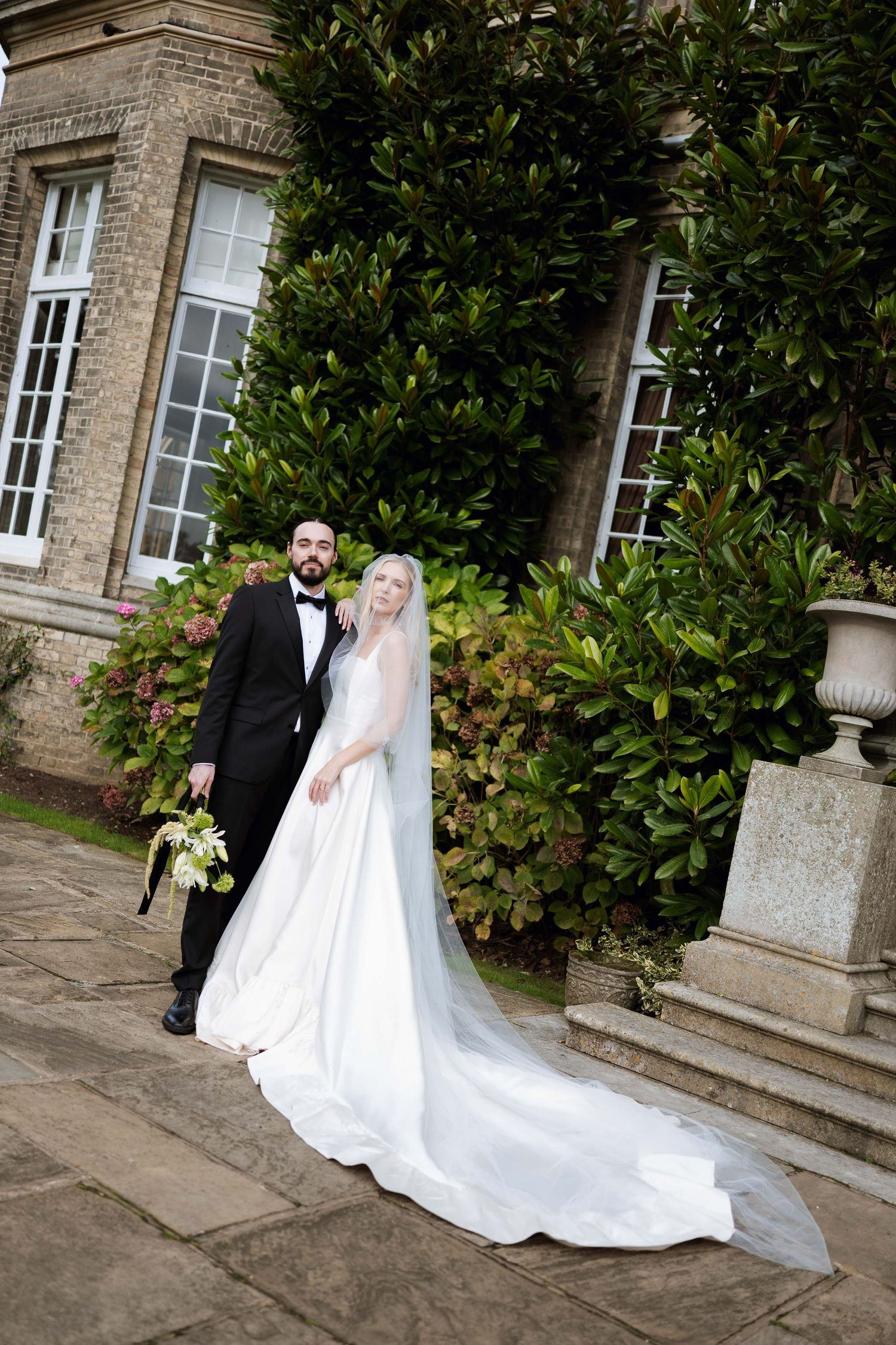 A bride and groom pose elegantly on stone steps outside a historic building with lush greenery in the background.