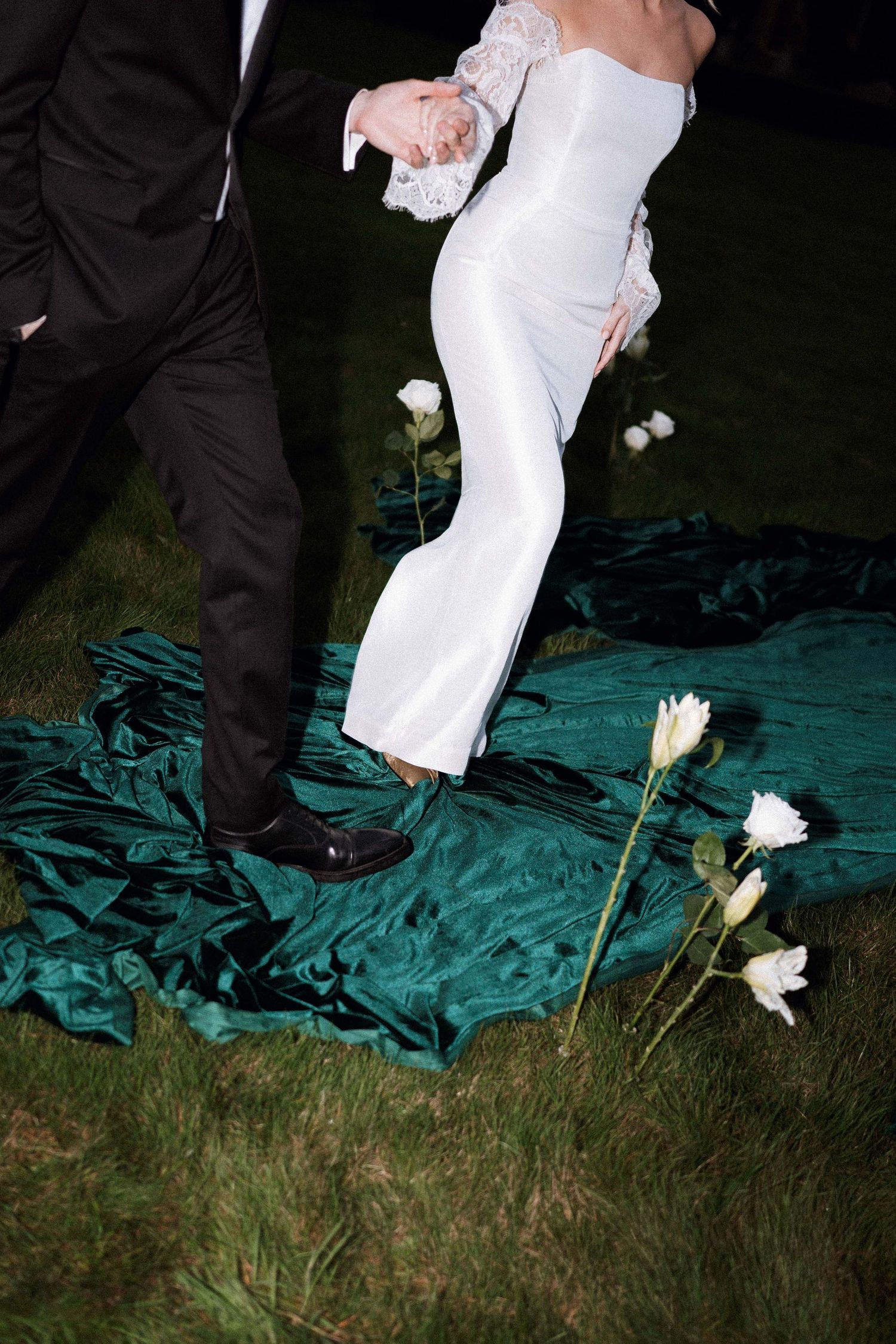 A couple in formal wedding attire dance together on a teal-colored surface with white flowers scattered nearby.