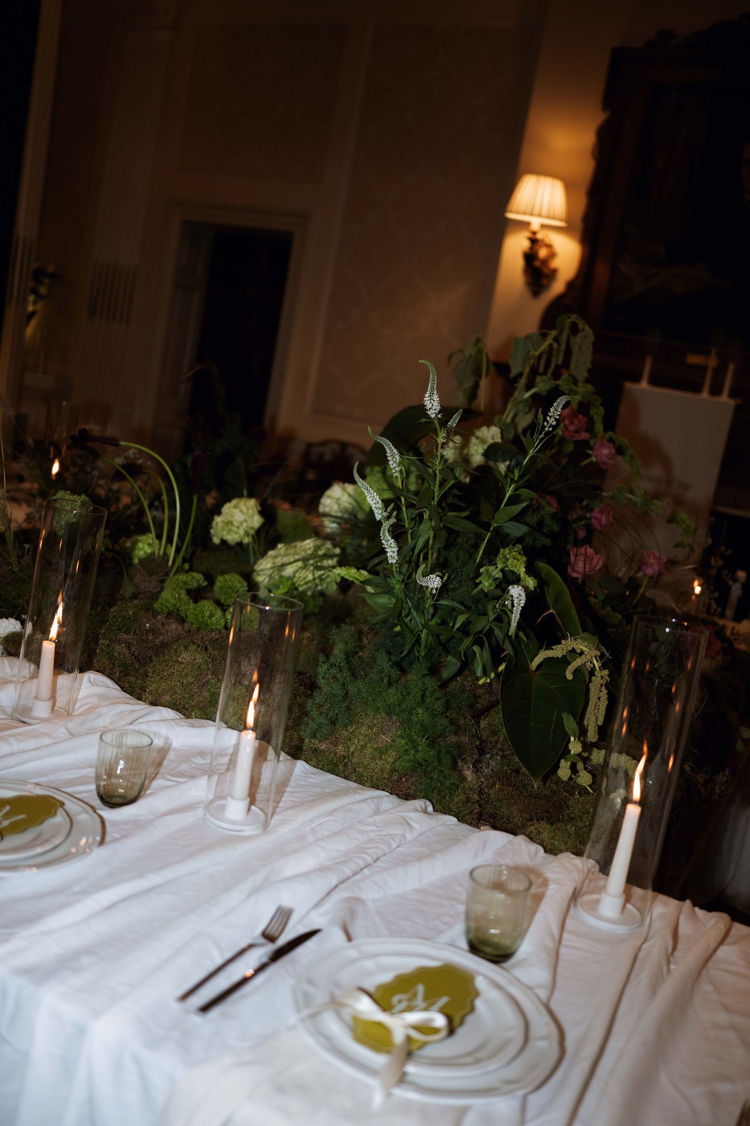 Elegant table setting with white linens, green foliage centerpiece, and glowing candles for an intimate dinner party.