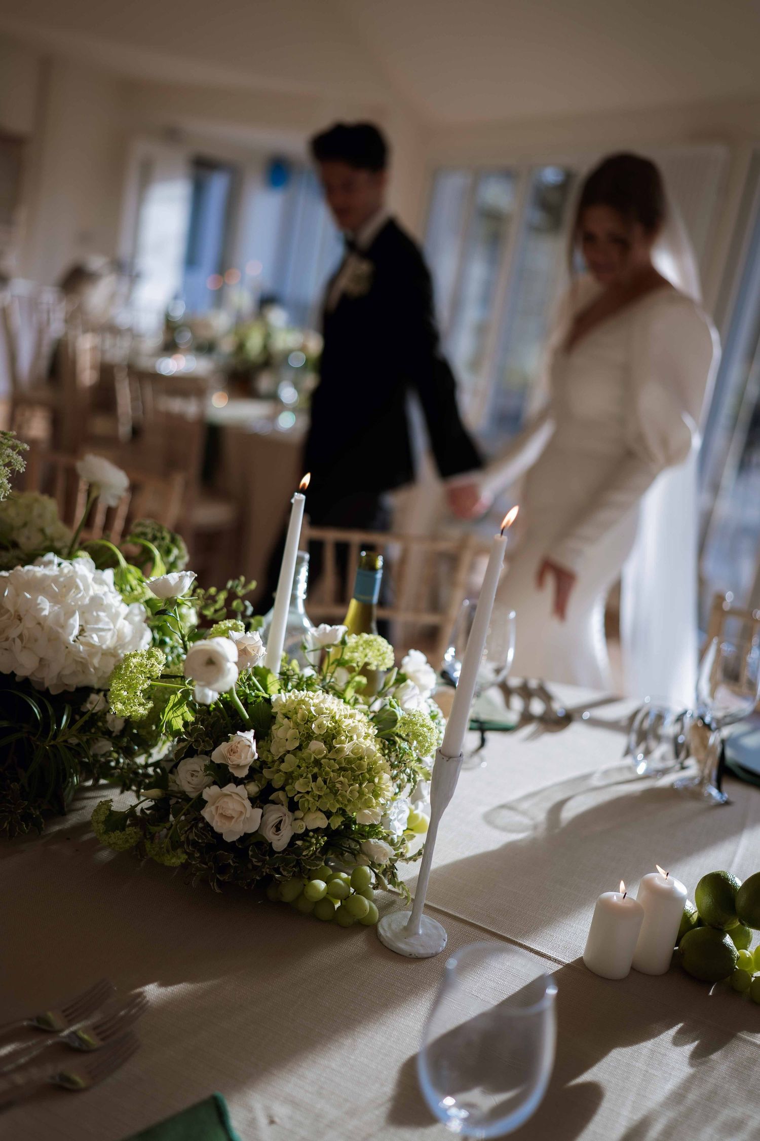 Elegant wedding table settings with white and green floral centerpieces and candles in a bright indoor venue.