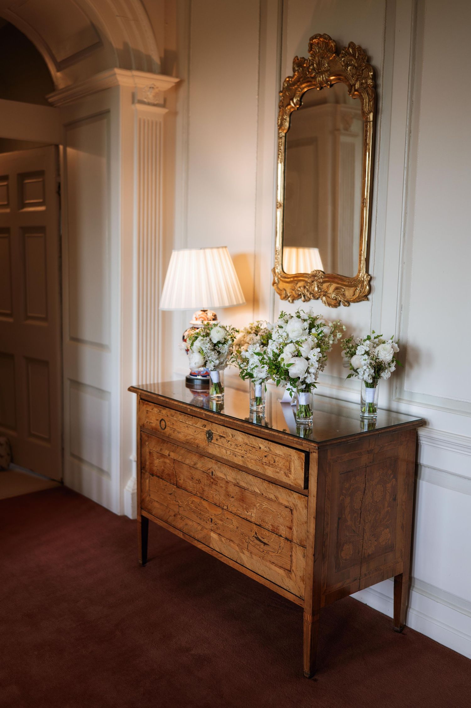 Elegant wooden dresser with ornate gold mirror and white floral arrangement in classic room with paneled walls.