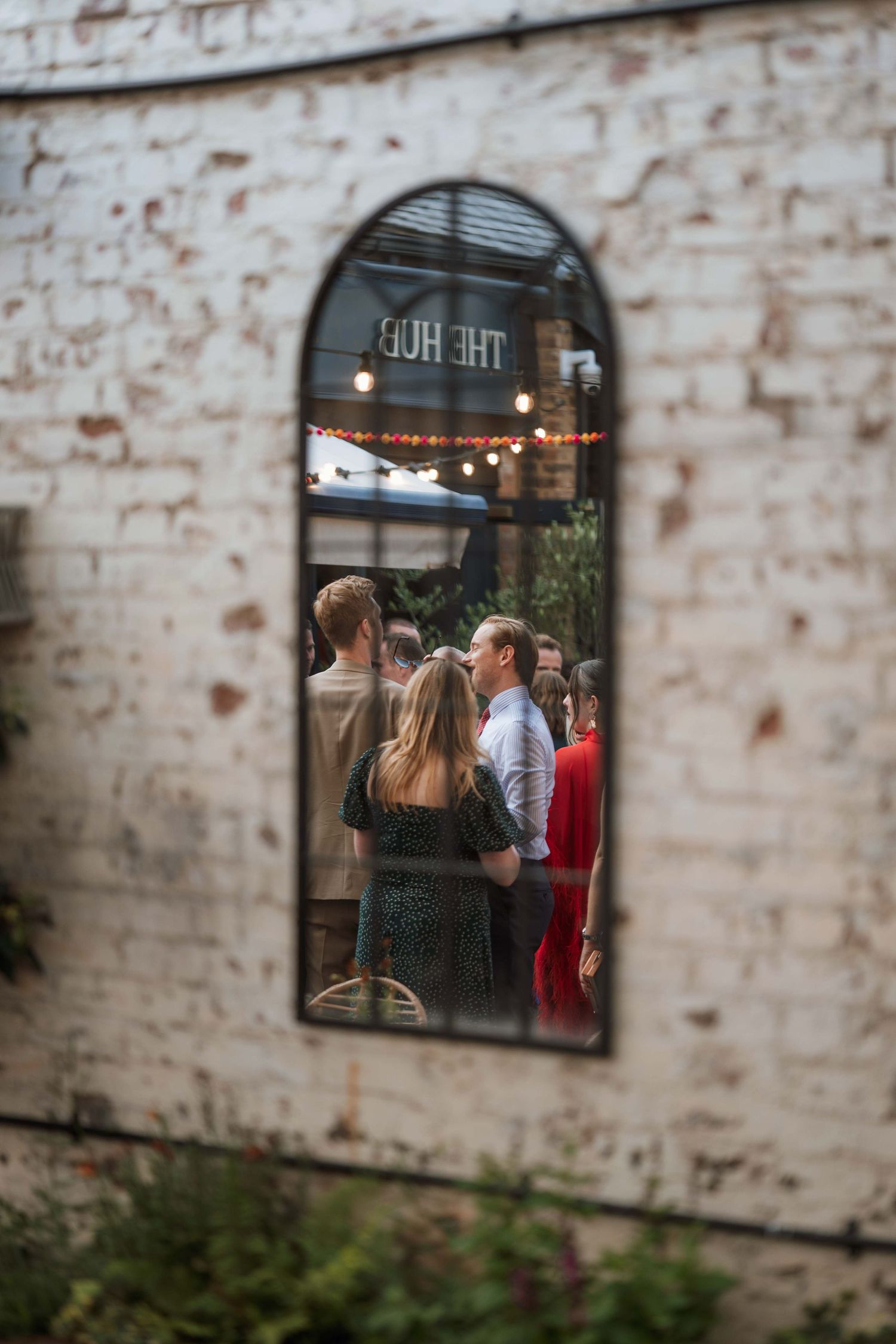 A series of windows in a rustic brick wall show glimpses of a cozy interior with string lights and people inside.