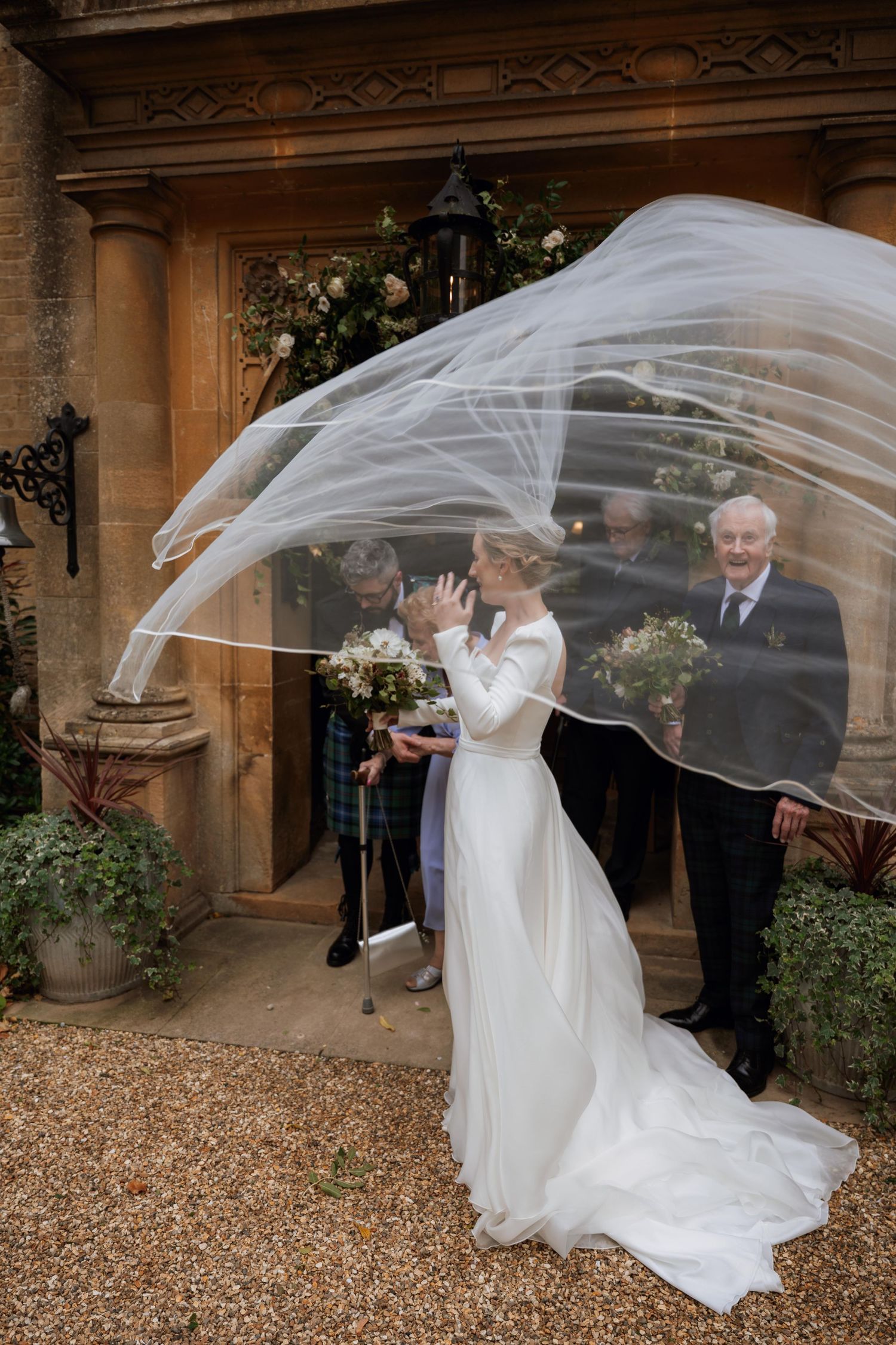 A flowing white wedding veil catches the wind dramatically on a gravel path outside a historic stone building.