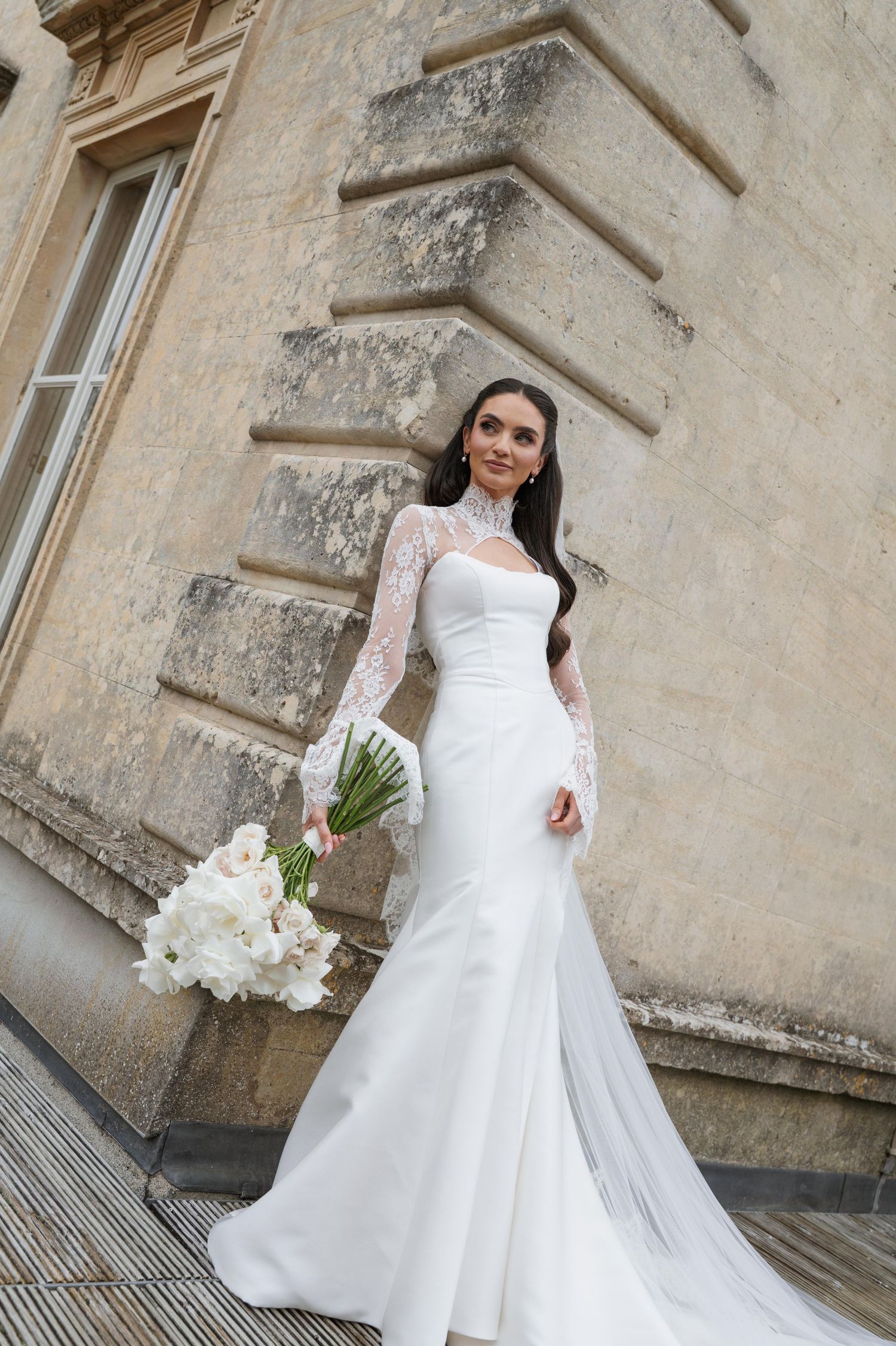 Bride in elegant white mermaid wedding gown with lace sleeves holding floral bouquet beside a stone building.