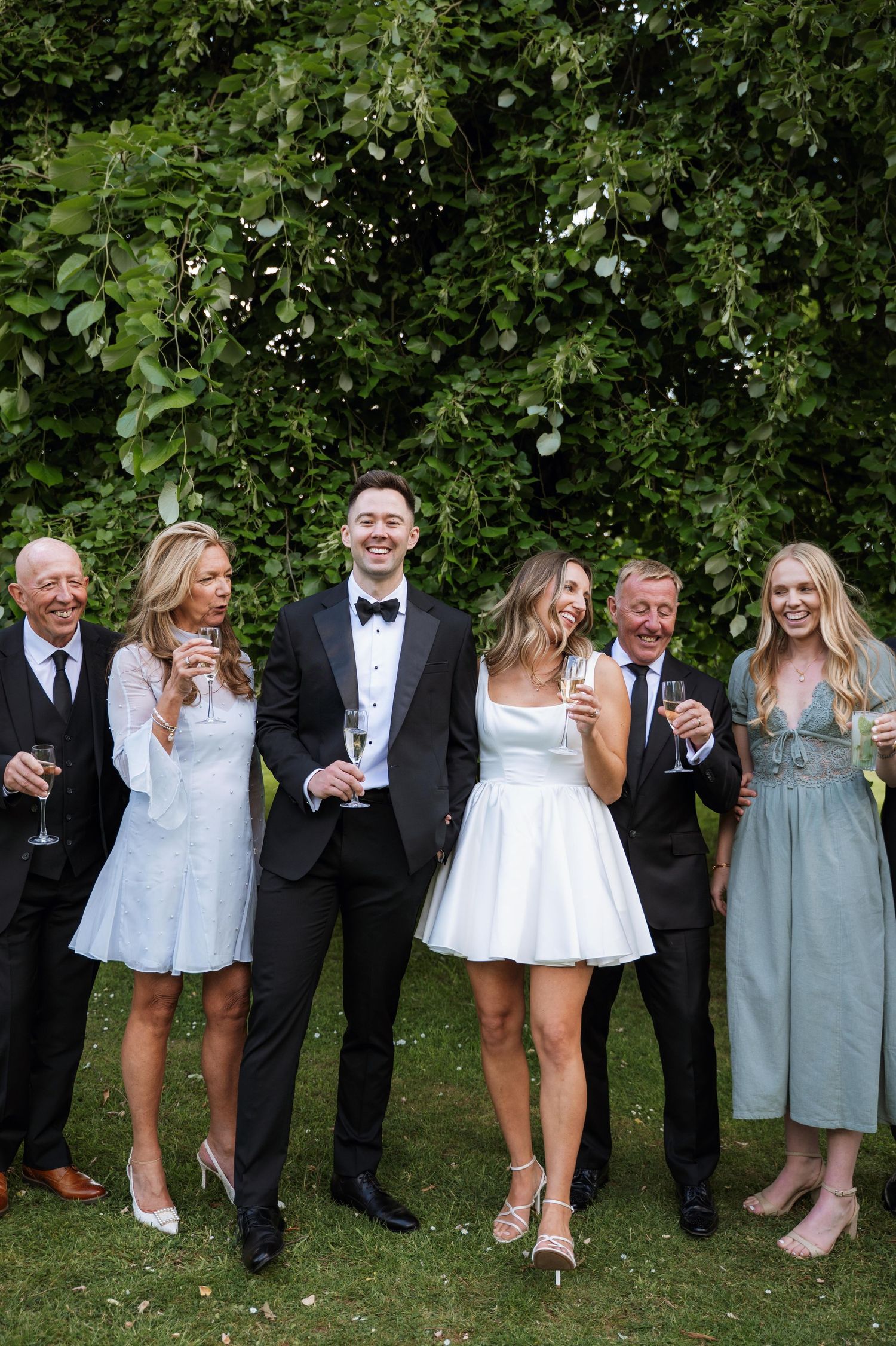 Wedding party members in formal attire stand in a row showing off their shoes against a green garden backdrop.