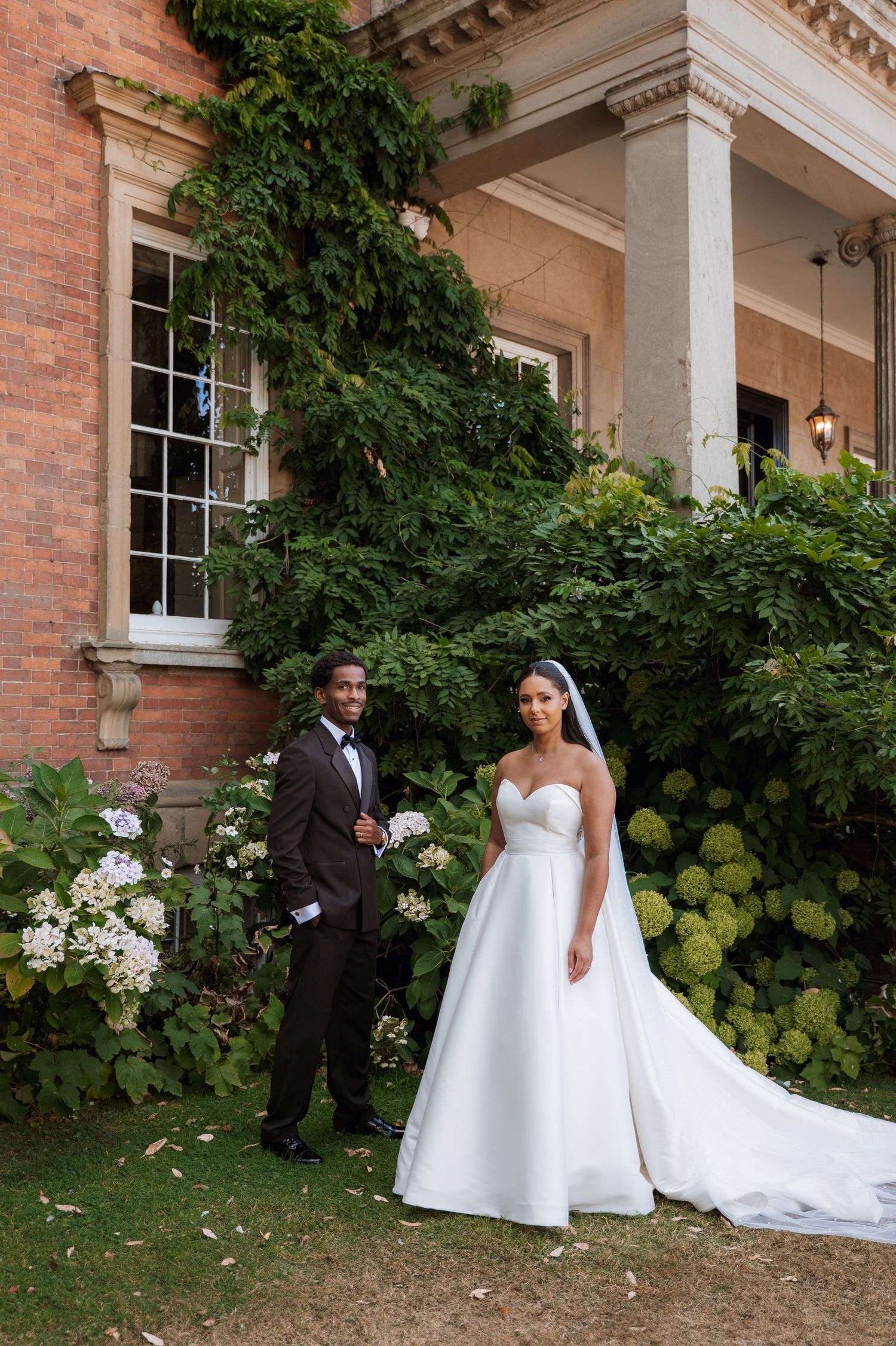 A couple in wedding attire poses together in front of a historic brick building with lush green ivy and white flowers.