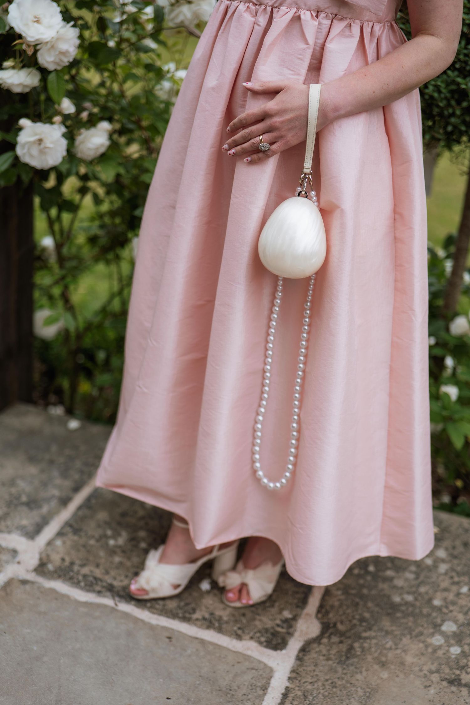 Woman in pink dress holding white pearl handbag accessory standing near white roses in garden setting.