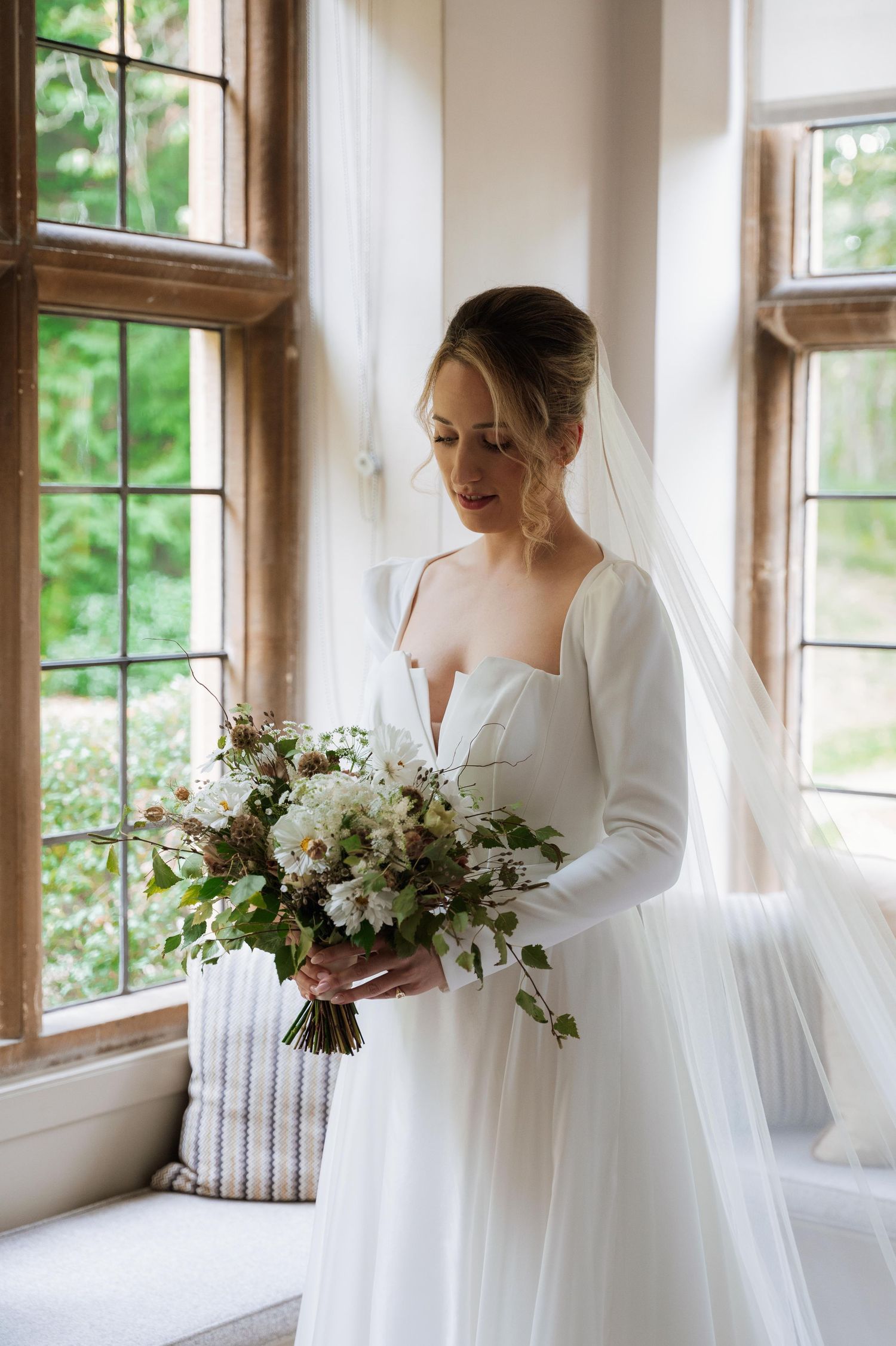 A bride in a flowing white dress stands by a window holding a delicate white and green floral bouquet.