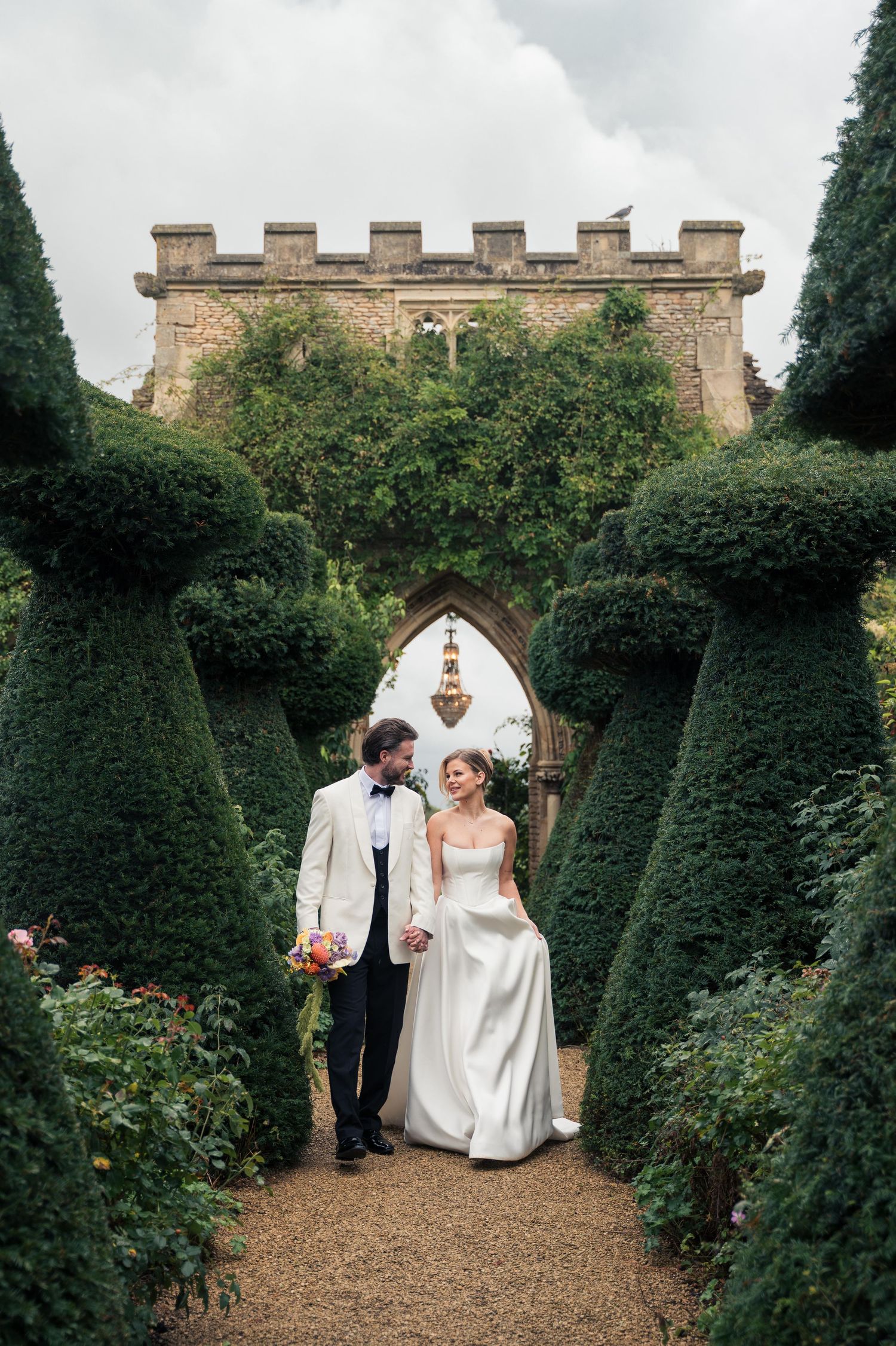 A wedding couple walks along a garden path lined with manicured topiary trees and stone walls at the elegant estate Euridge Manor.