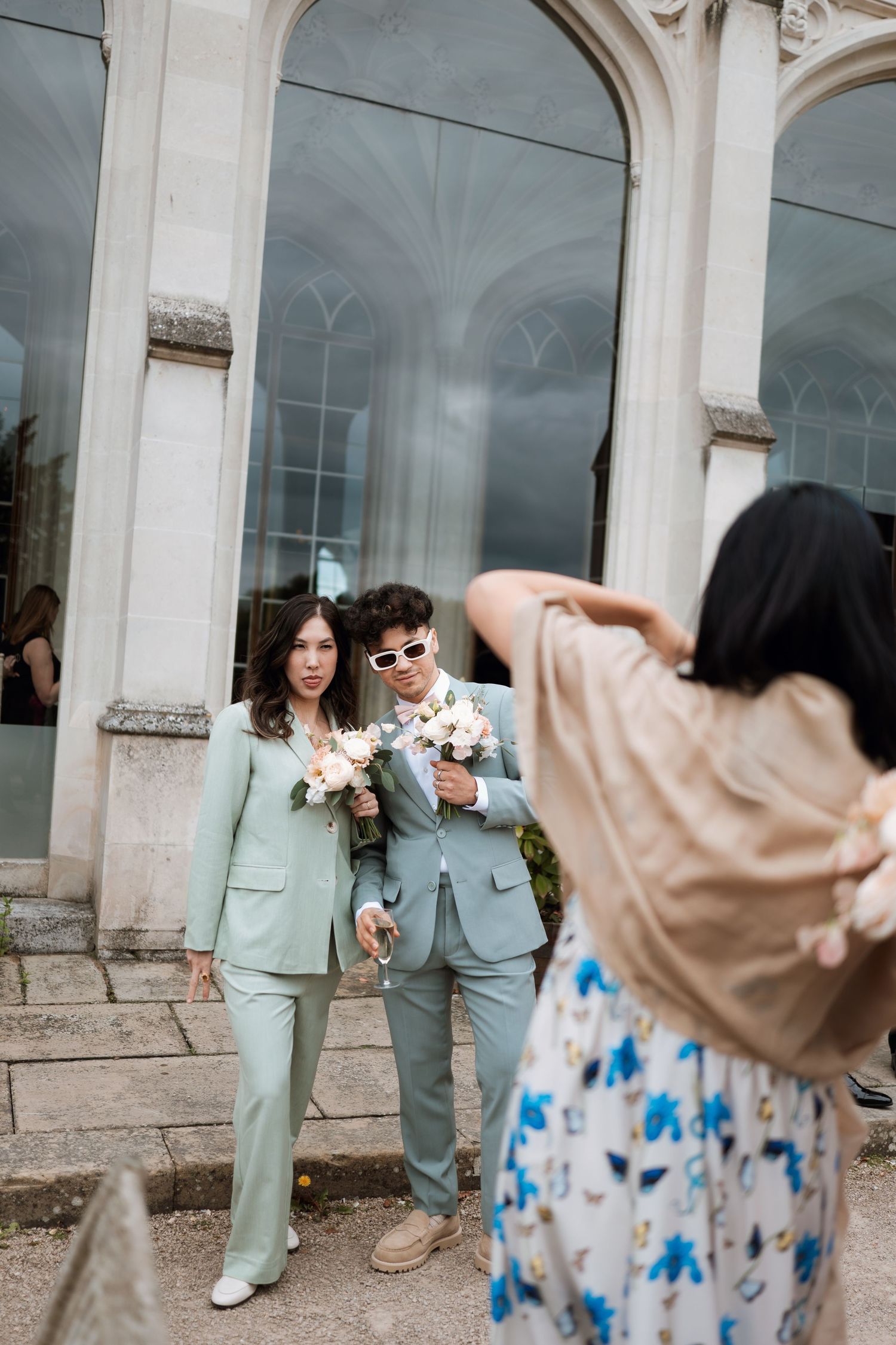 Two people in matching mint green suits hold bouquets while being photographed outside an elegant arched building entrance.