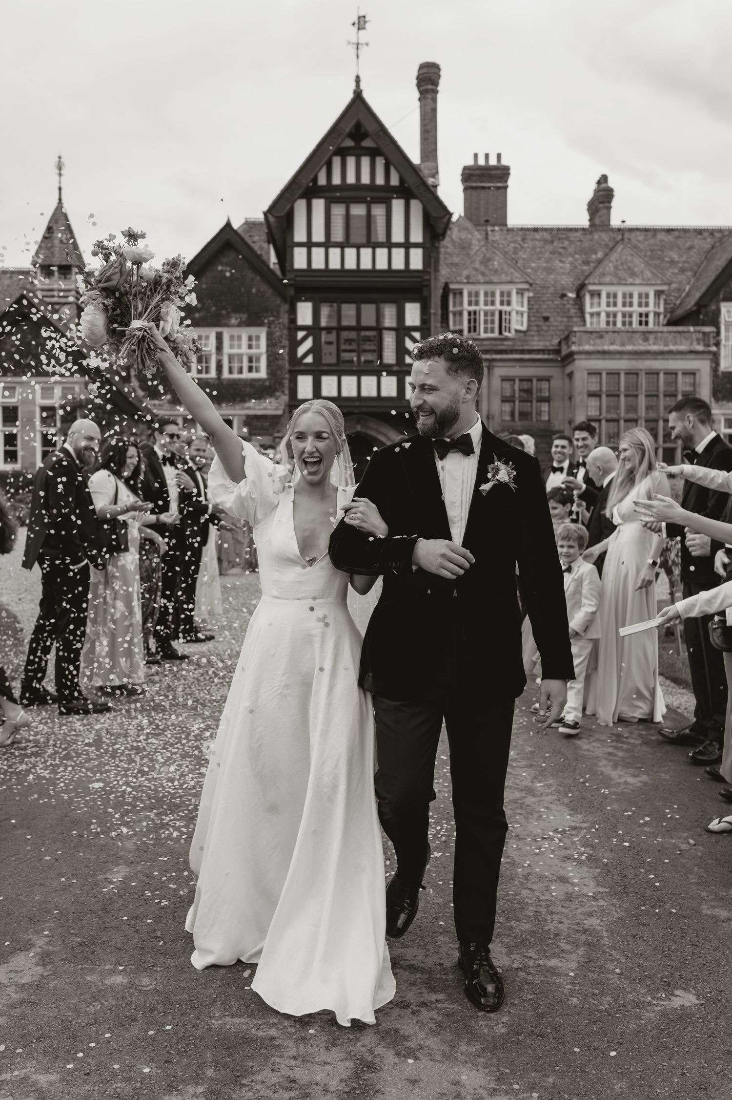 Newlyweds walk through a shower of confetti outside a Tudor-style building while guests celebrate their union.