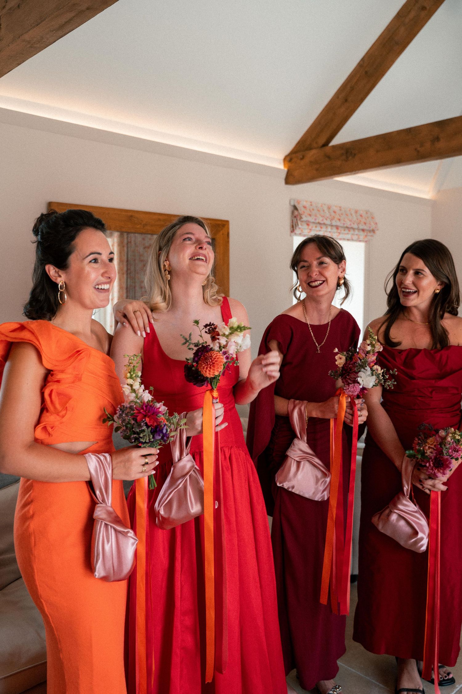 Wedding party in orange and burgundy dresses laughing together while holding bouquets in a rustic venue.