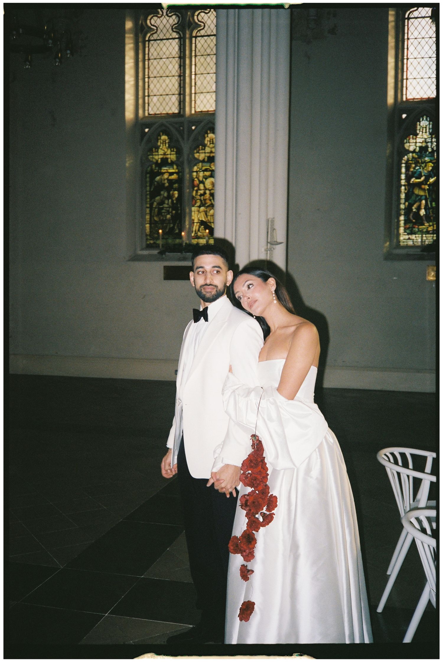 A couple in white wedding attire embraces in a church setting with stained glass windows and folding chairs.