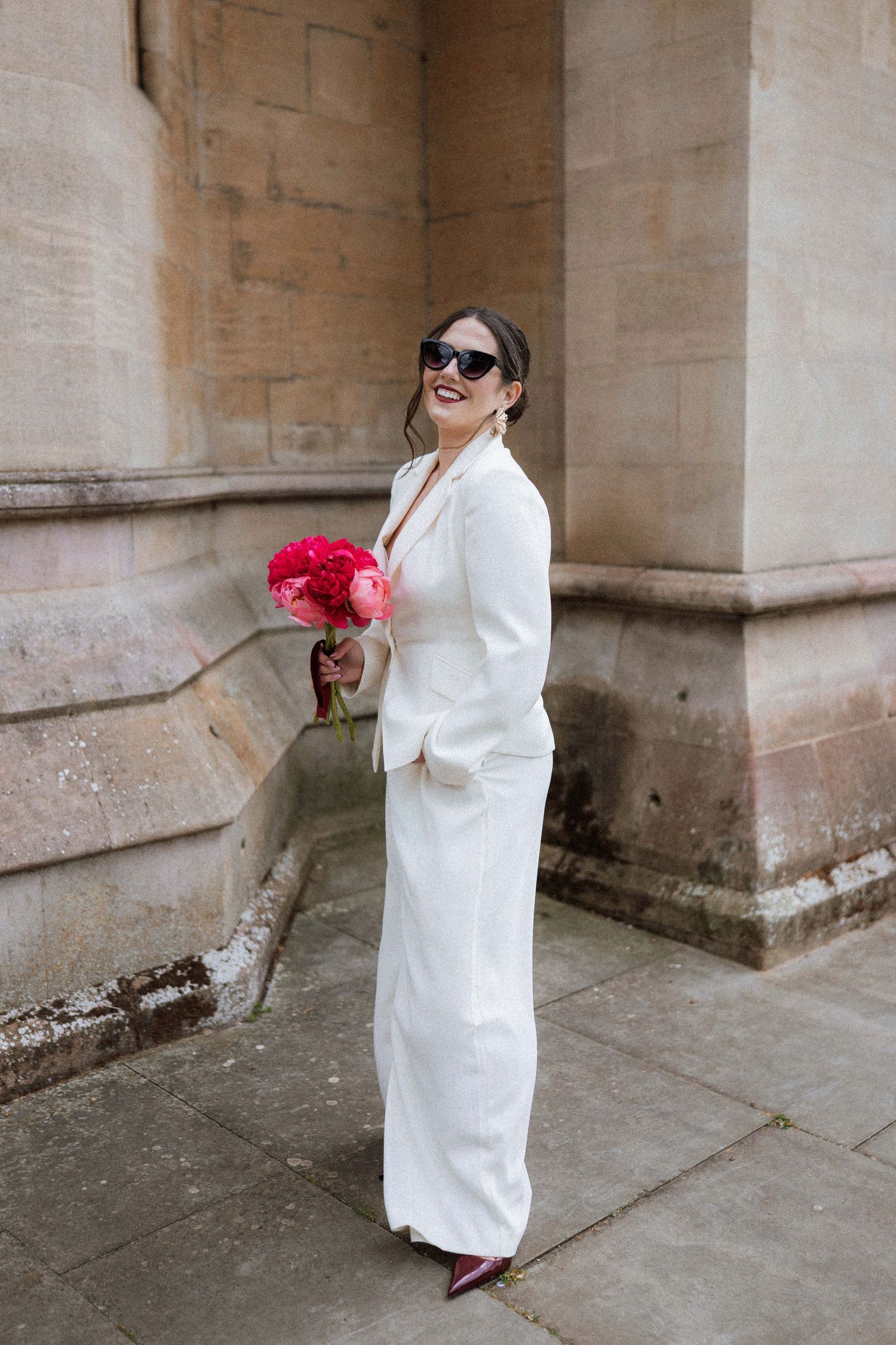 A person in a white coat and sunglasses poses with red flowers against stone columns in an outdoor setting.