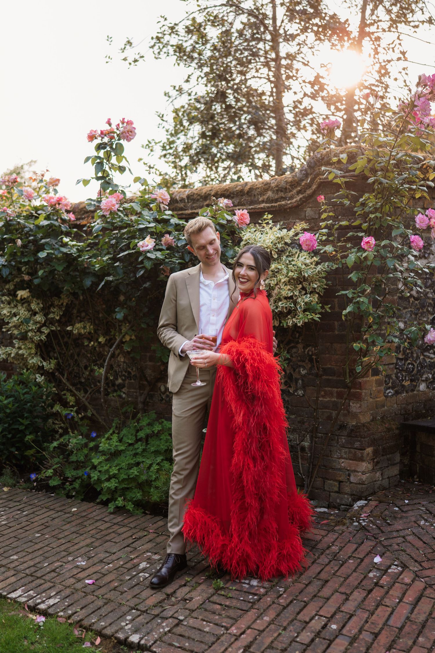 A couple poses for photos in a garden setting with blooming roses, featuring a striking red feathered gown.