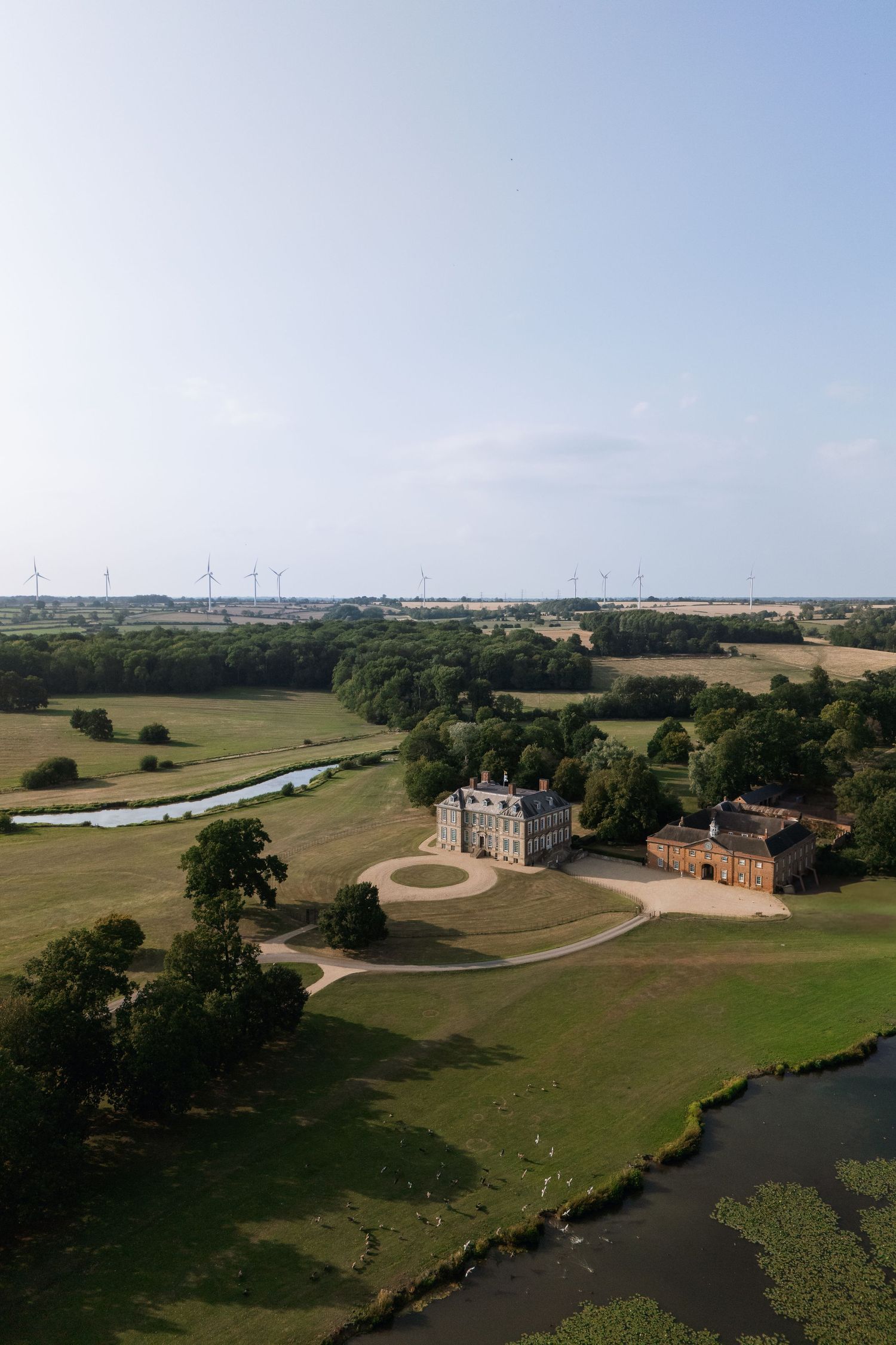 Aerial panoramic view of historic manor house buildings nestled among green fields along winding river valley.