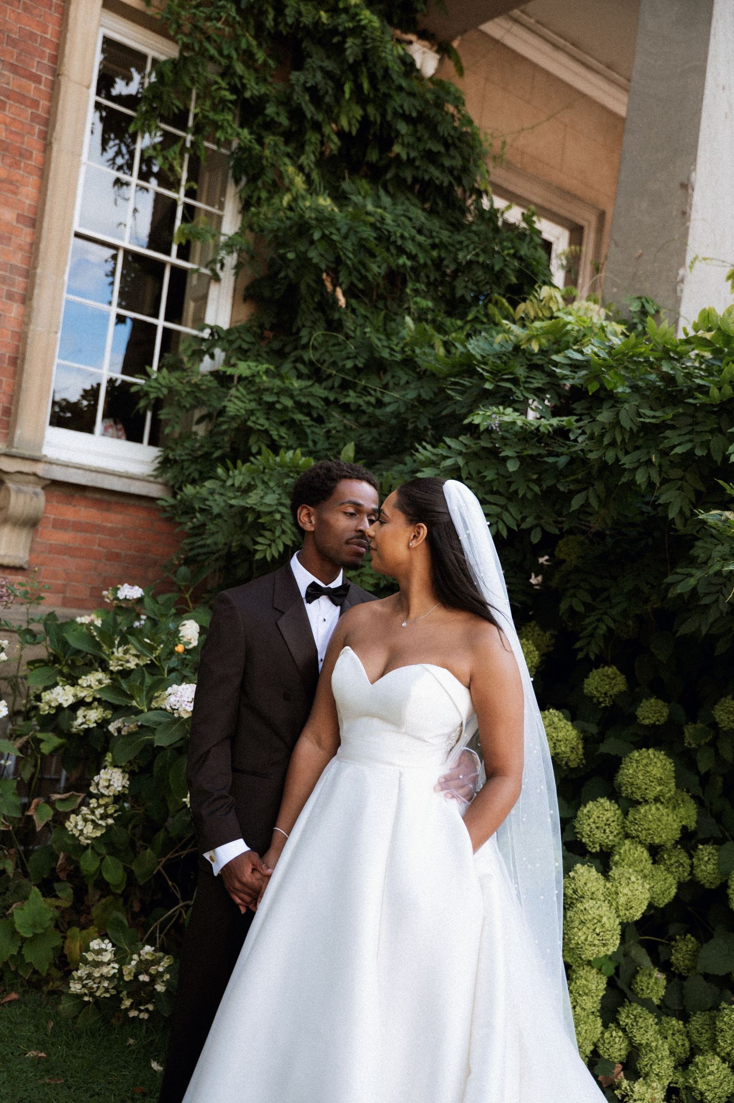 A happy couple in wedding attire share intimate moments in front of an elegant brick building with climbing vines.