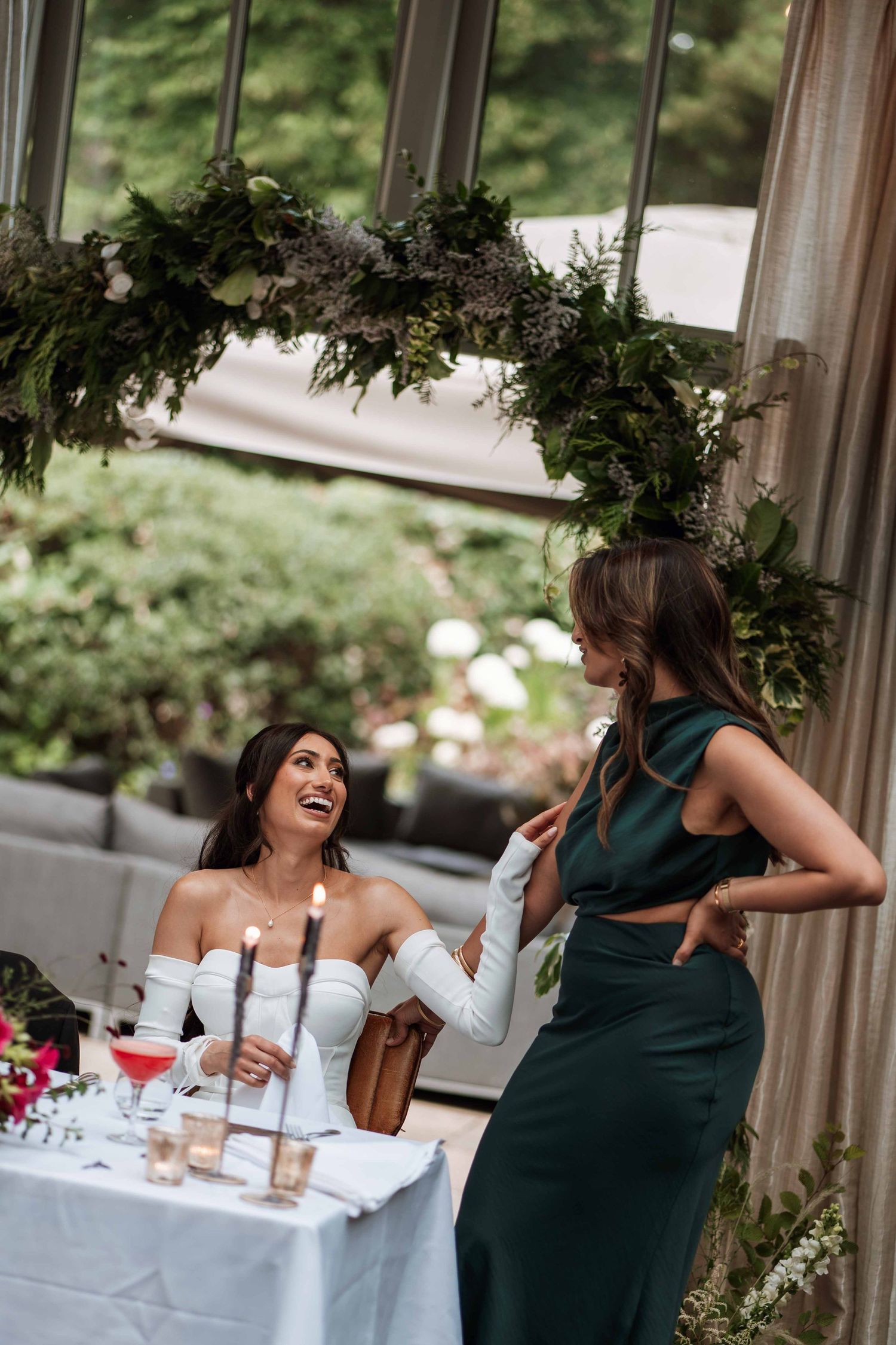 Two women sharing a joyful moment at an elegant reception table decorated with greenery garlands and white linens.