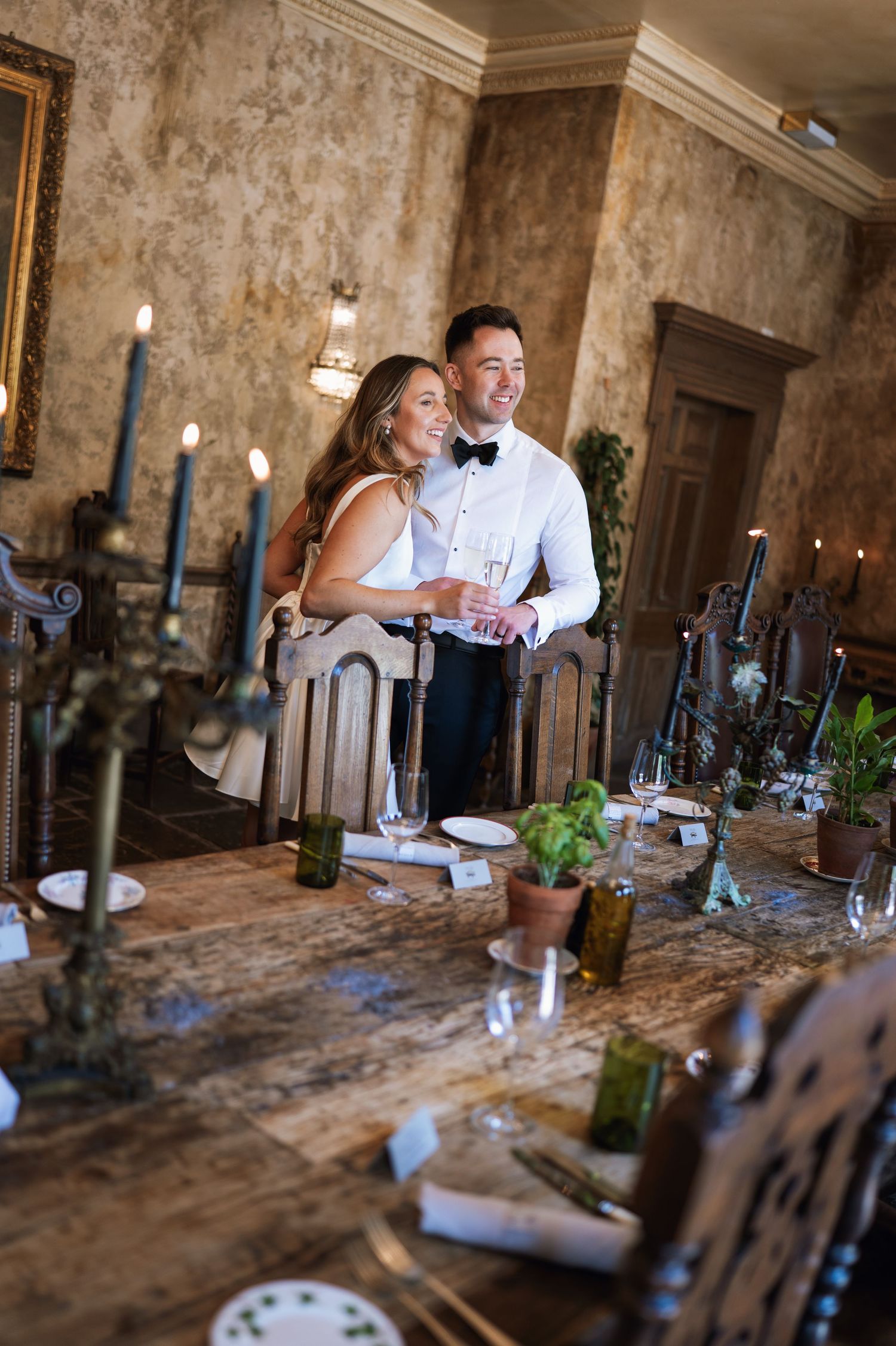A couple laughs together at an elegant dinner table with candles and rustic decor in a stone-walled room.