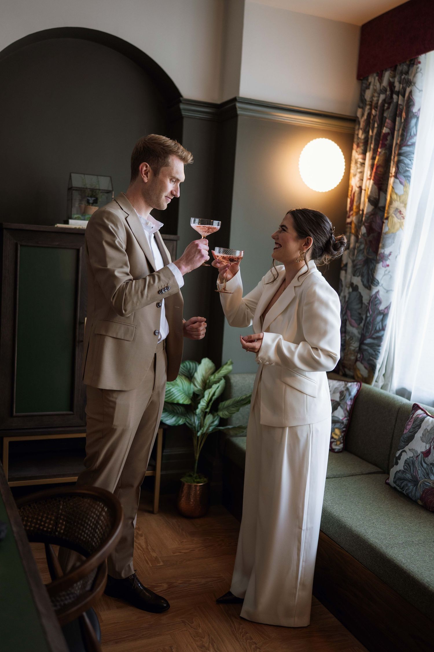 Two people in elegant attire enjoying cocktails while having a conversation in a cozy vintage-style room.