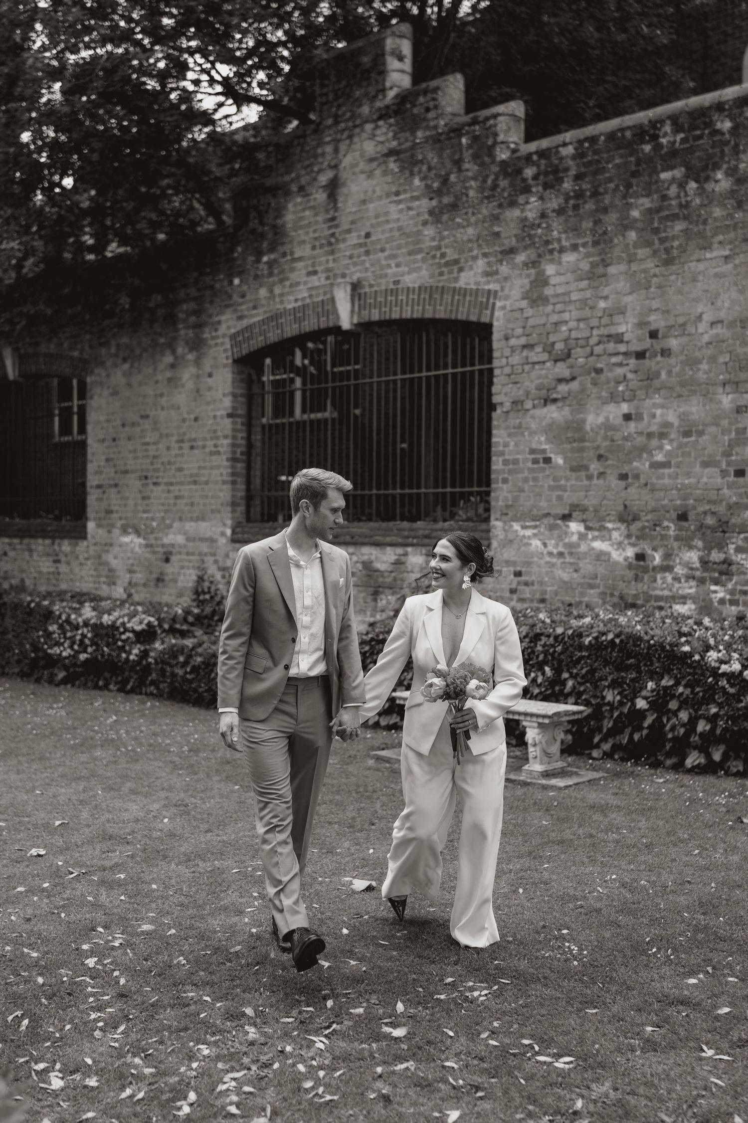 A couple in formal wedding attire walks through a garden with brick walls and scattered leaves on the ground.