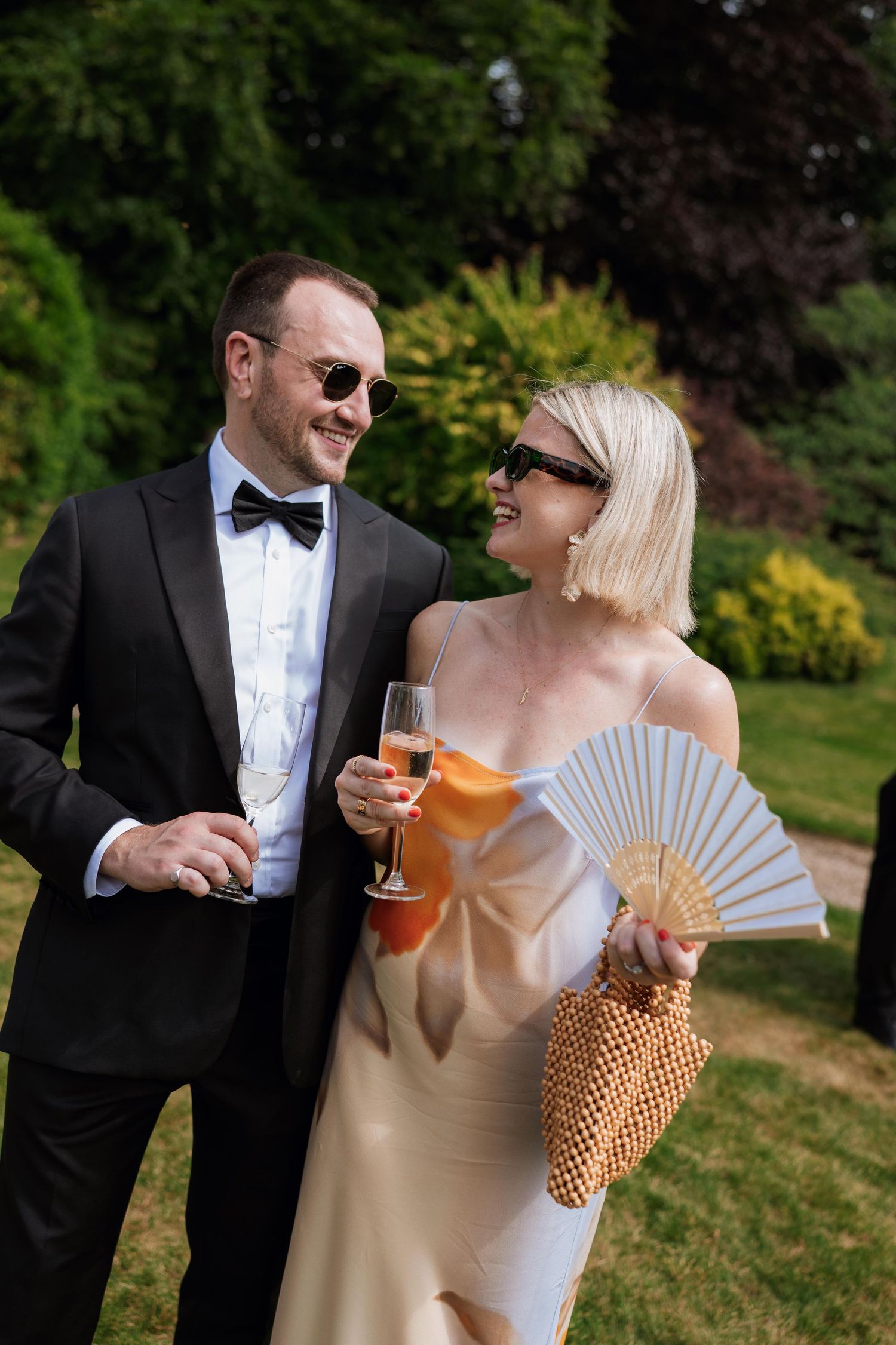 Couple in formal attire sharing drinks and laughing together while holding decorative fans in an outdoor garden setting.