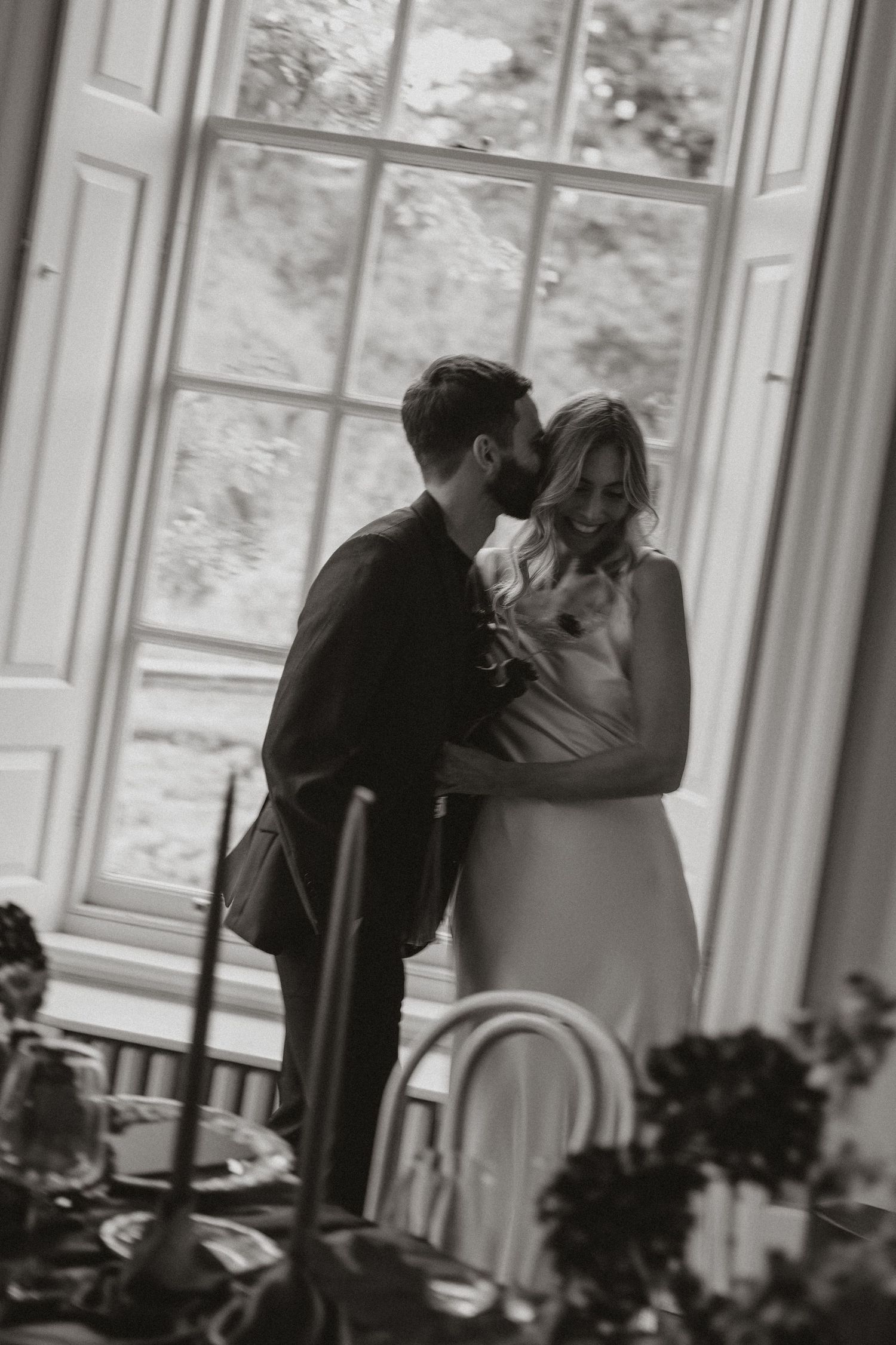 Black and white photo showing a romantic moment between two people in an elegant room with large windows.