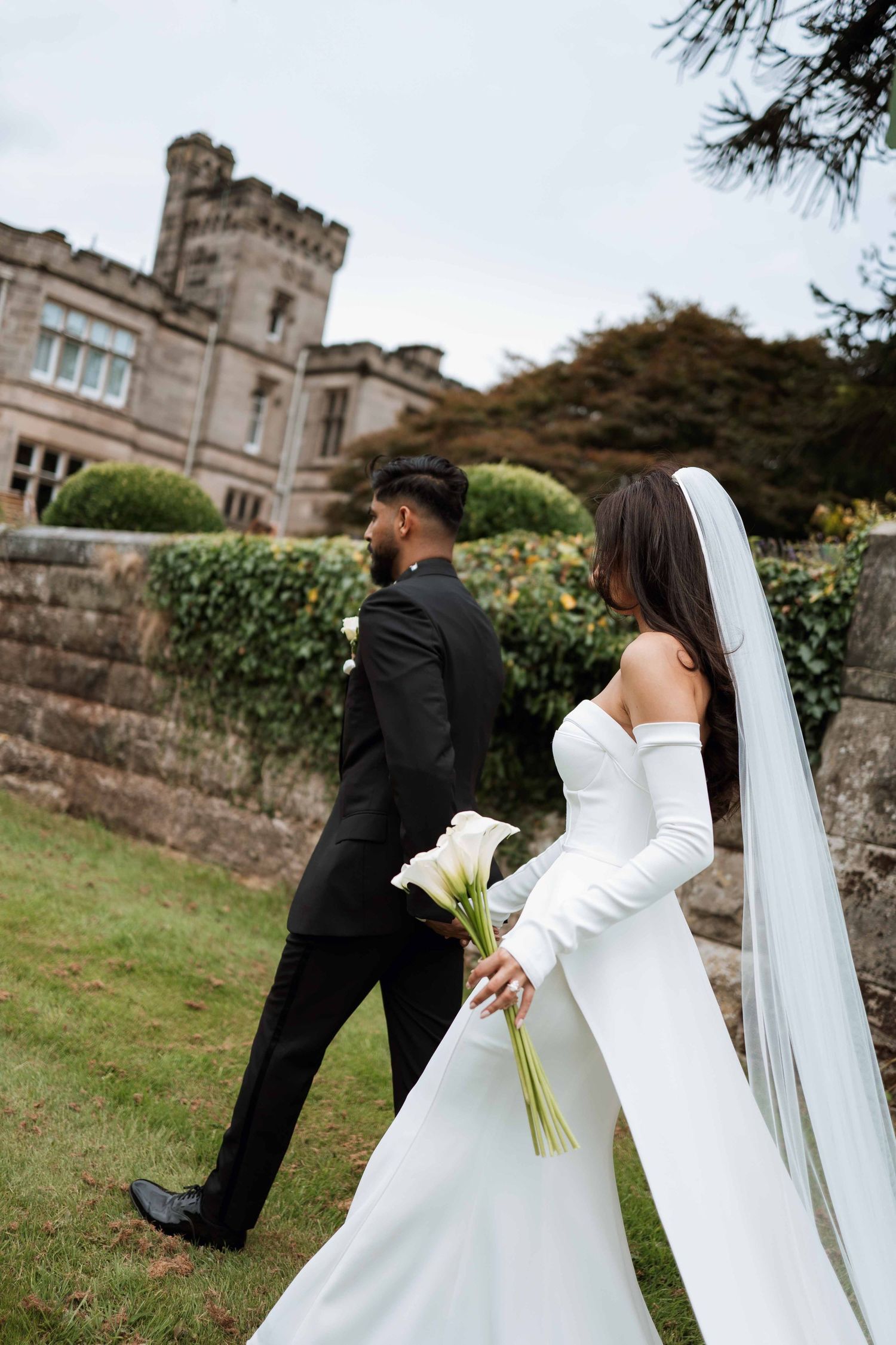 A bride in a white off-shoulder gown with long veil holds calla lilies near a castle-like building with stone walls.