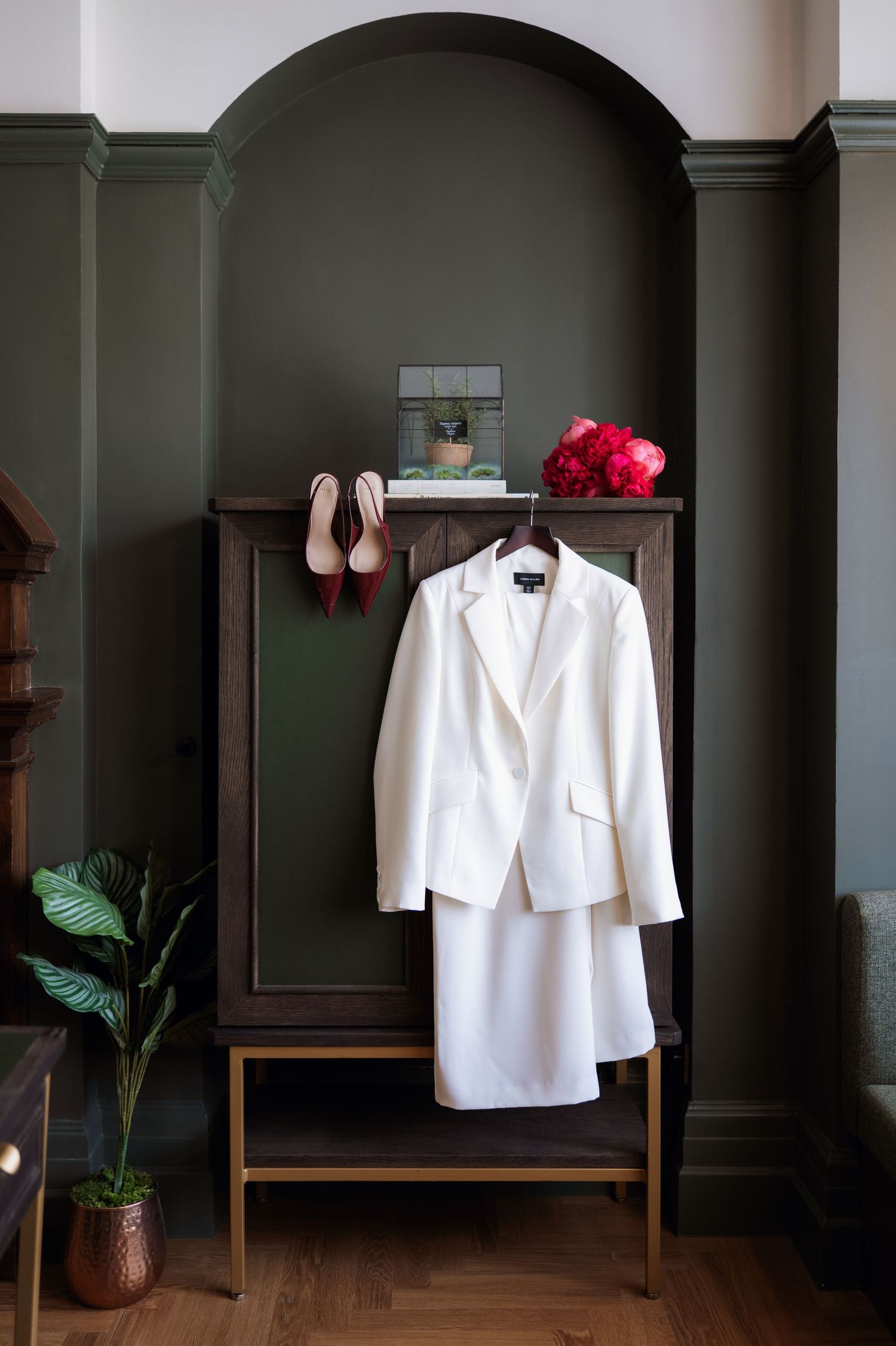 White formal jacket hangs on a wooden rack against dark green walls with decorative pink flowers and potted plants.
