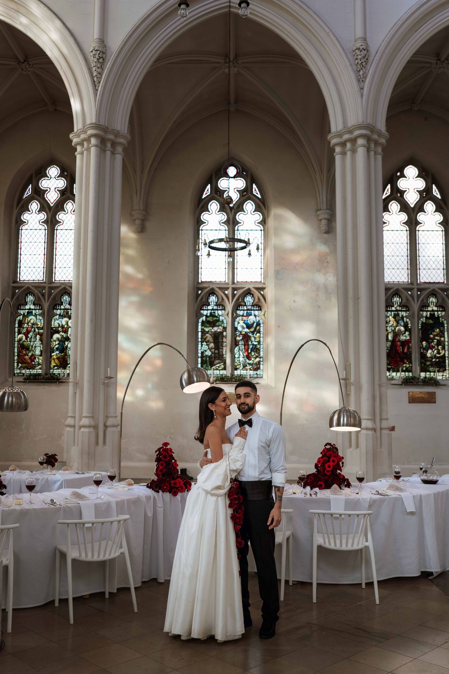 A romantic wedding reception setup in a historic chapel with gothic windows, white tables and elegant arched decorations.