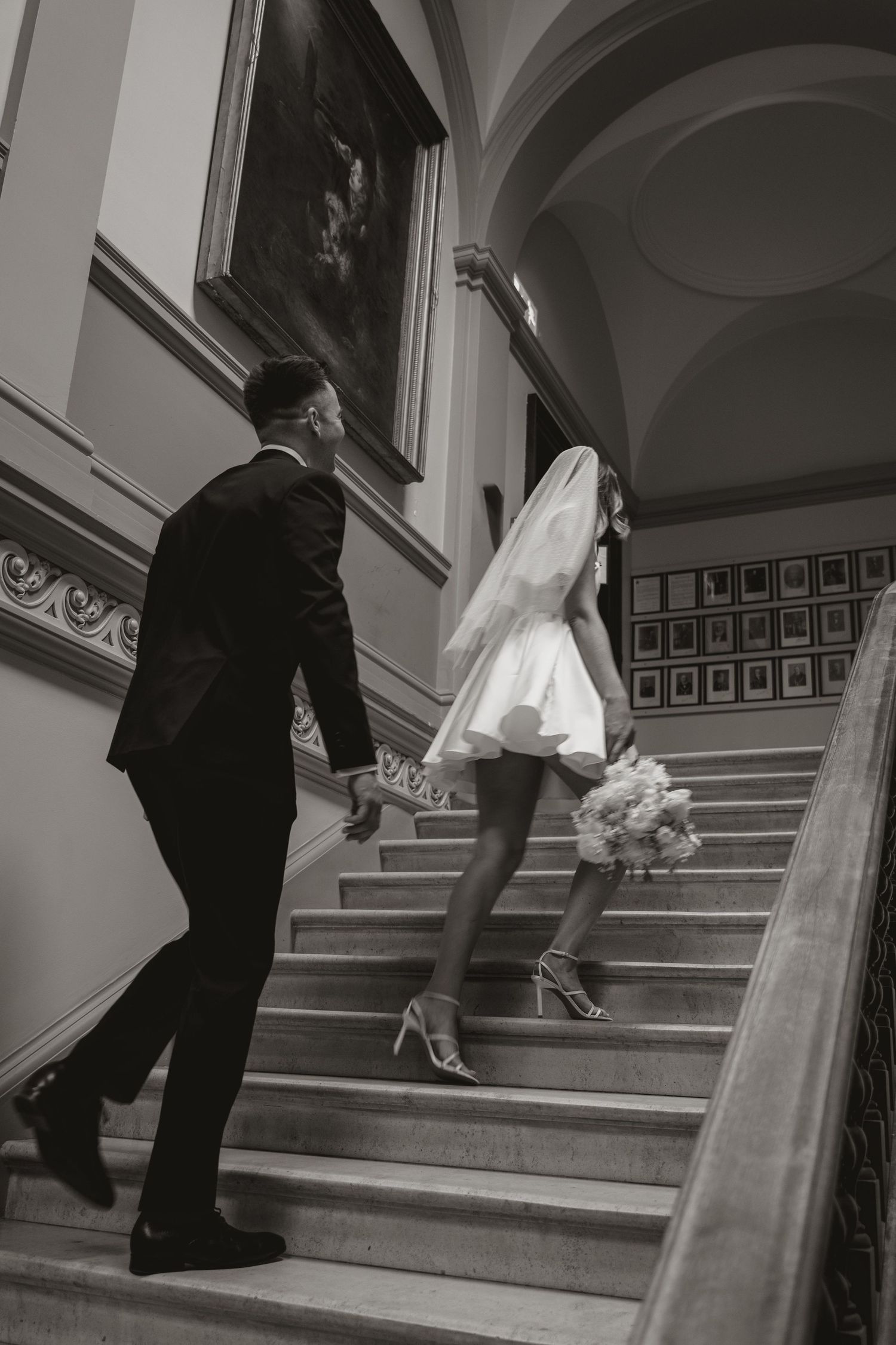 Black and white photo of newlyweds ascending elegant stairs, bride in short dress carrying bouquet with groom following.