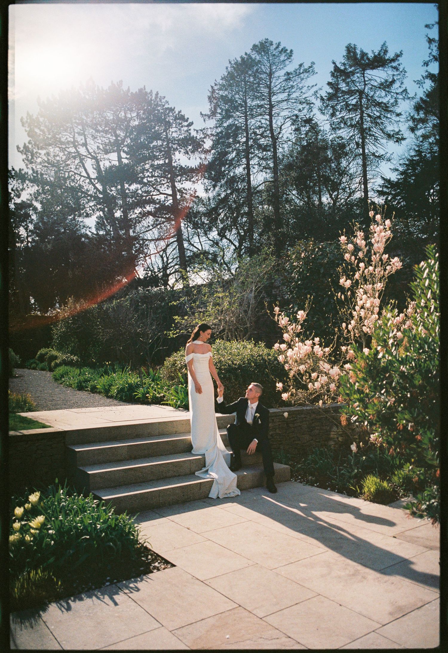 Bride in white gown stands on garden steps as groom kneels beside her, surrounded by lush greenery and blooming trees.