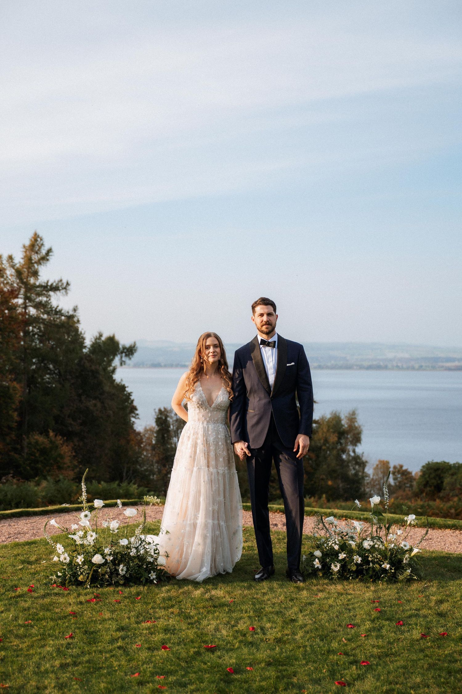 A couple in wedding attire stands on a grassy hillside overlooking a scenic bay with mountains in the background.