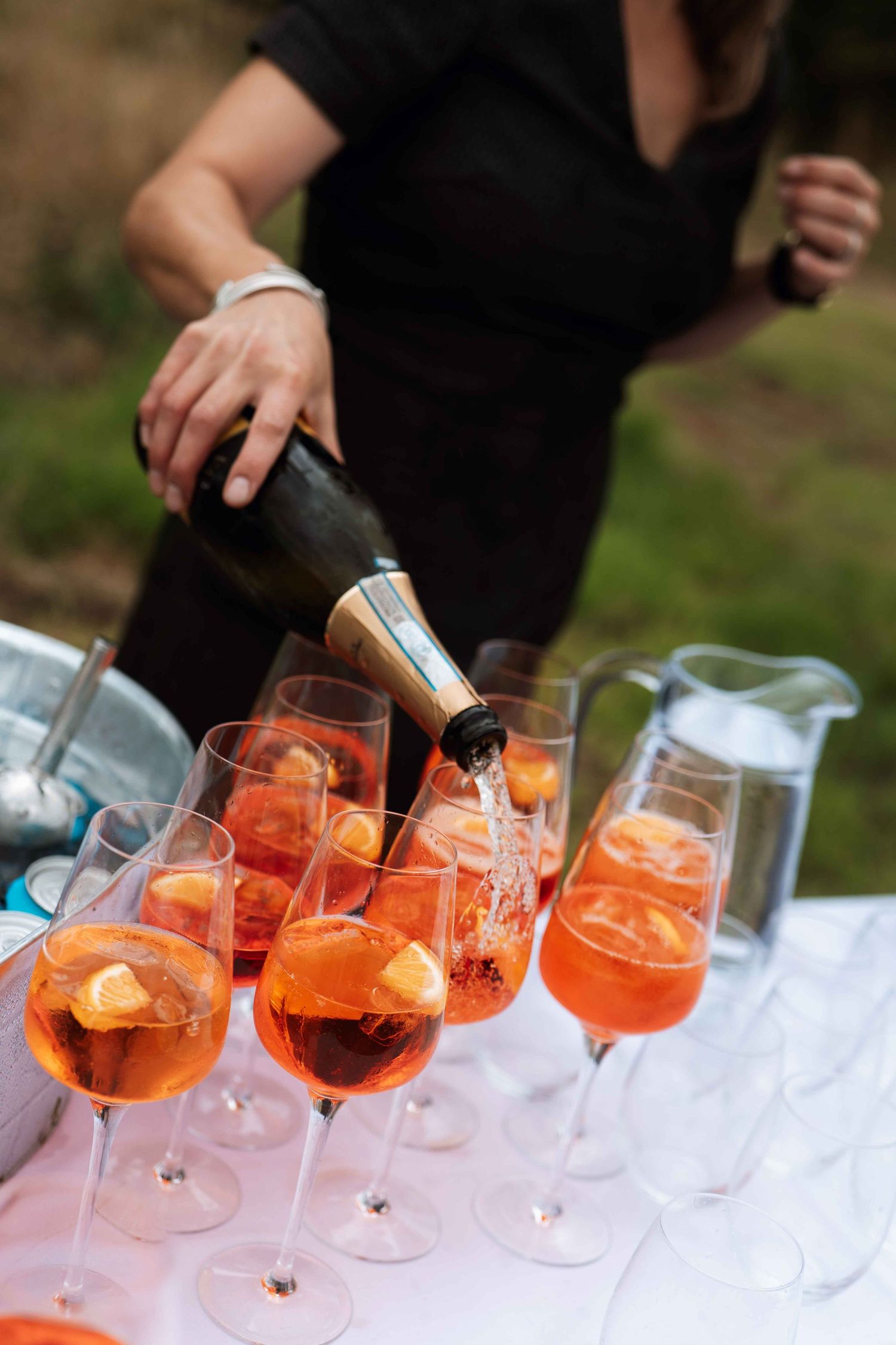 Hands pouring orange Aperol Spritz cocktails into multiple wine glasses arranged in a row on a white surface.