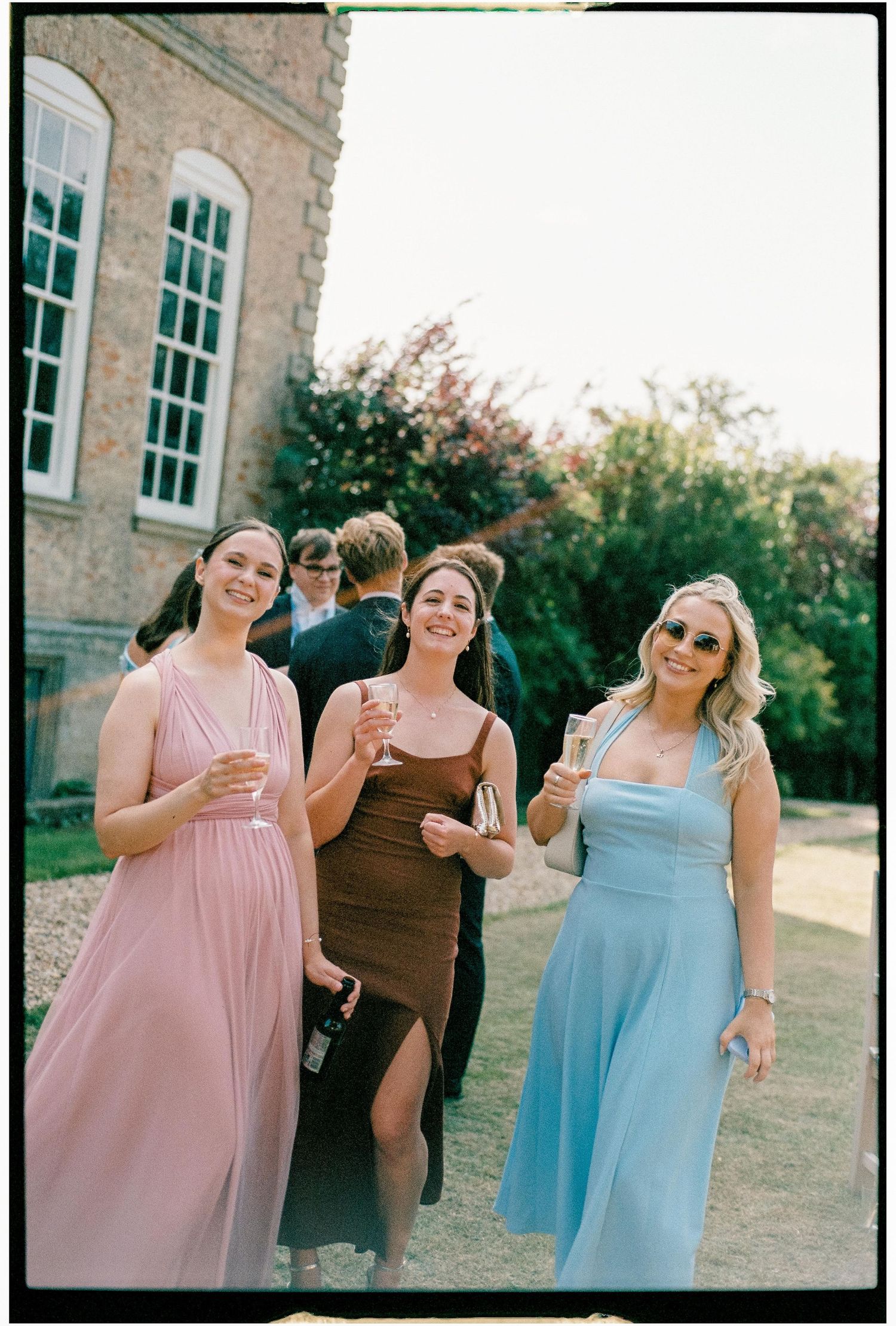 Three friends in pastel dresses share laughter and smiles while posing together outside a brick building.