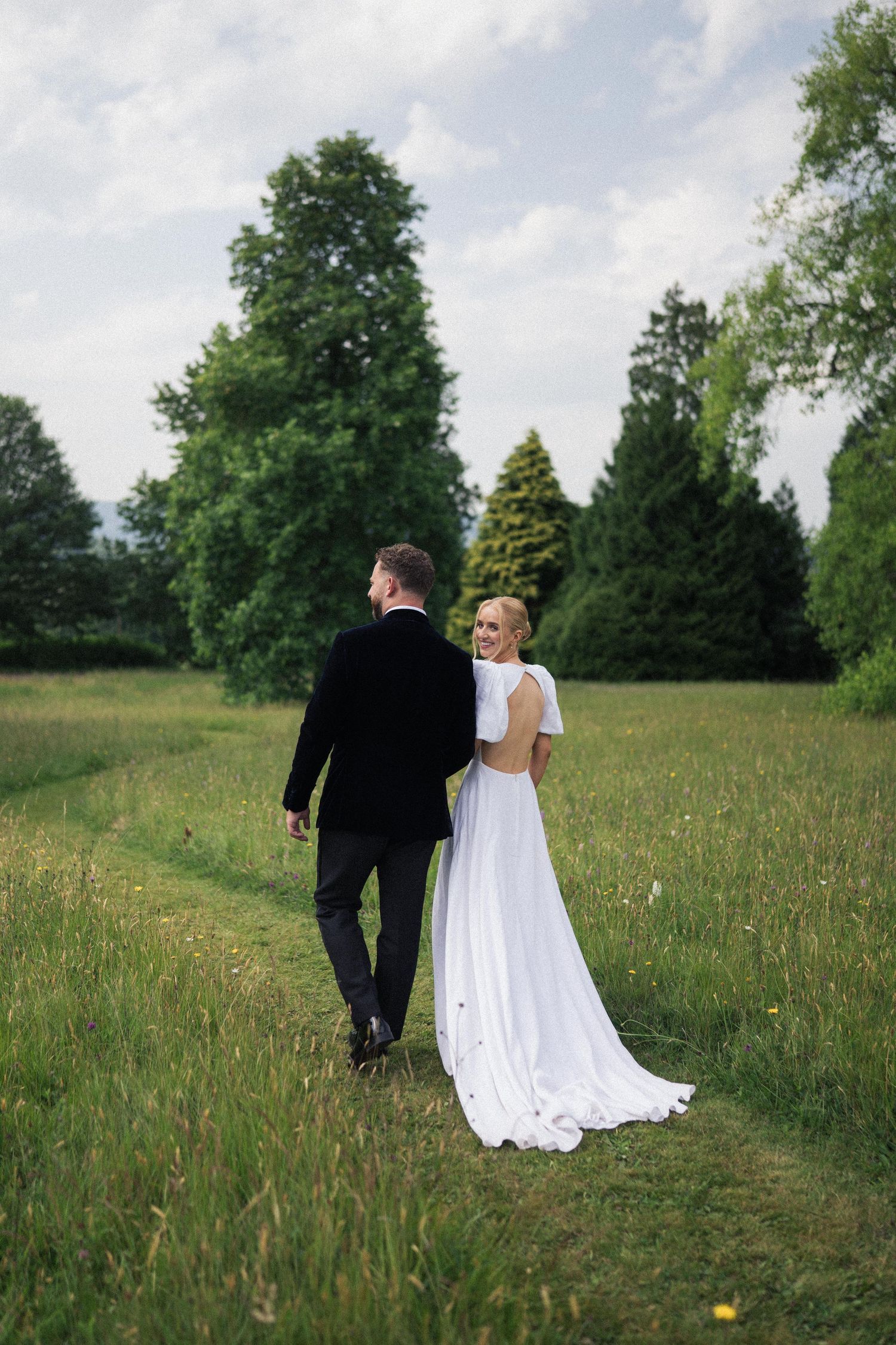 A couple in wedding attire walks through a lush green meadow on a summer day.