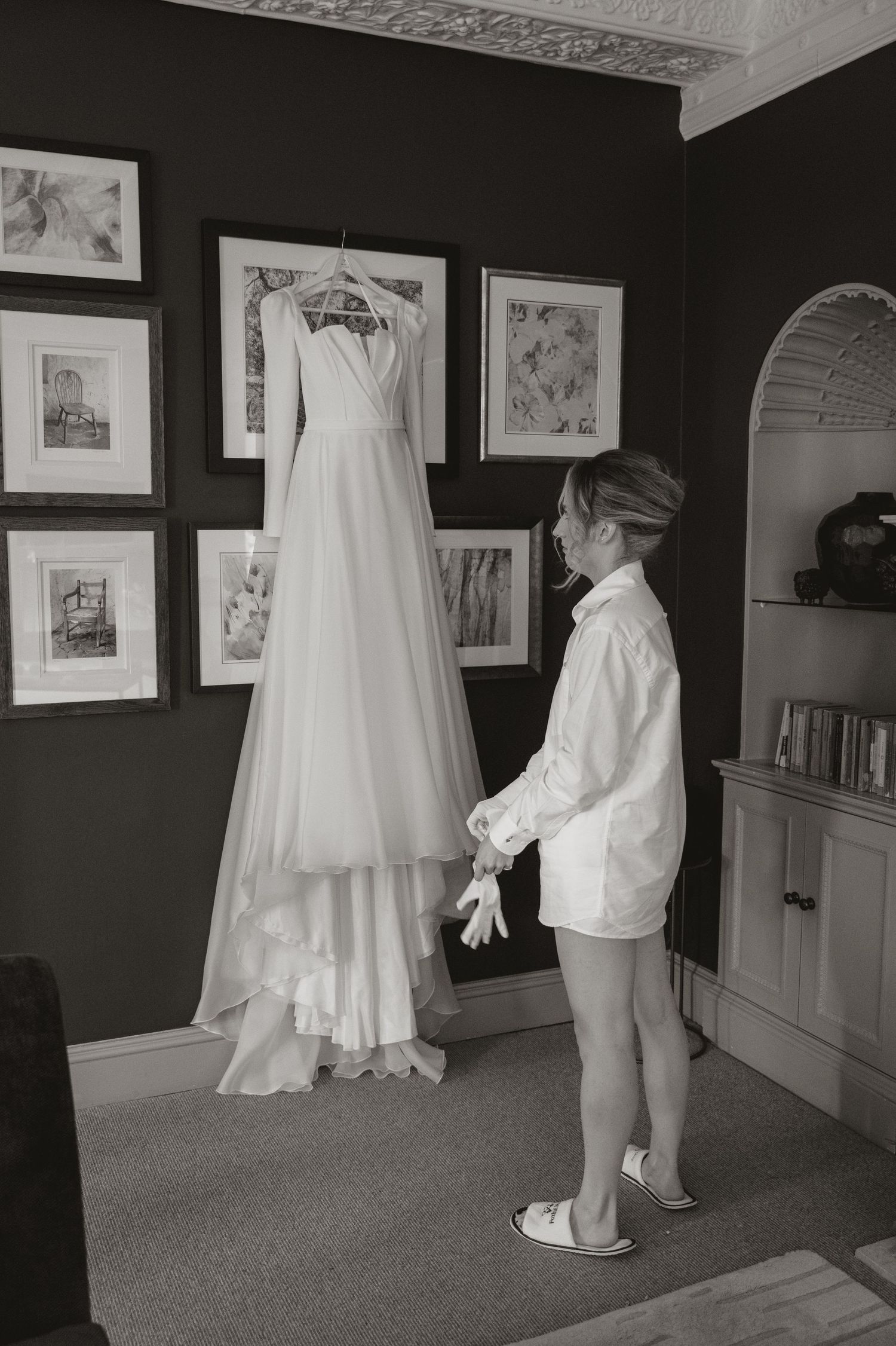 A young child in white shorts and shirt looks up at an elegant white wedding dress hanging on display in a room with framed art.
