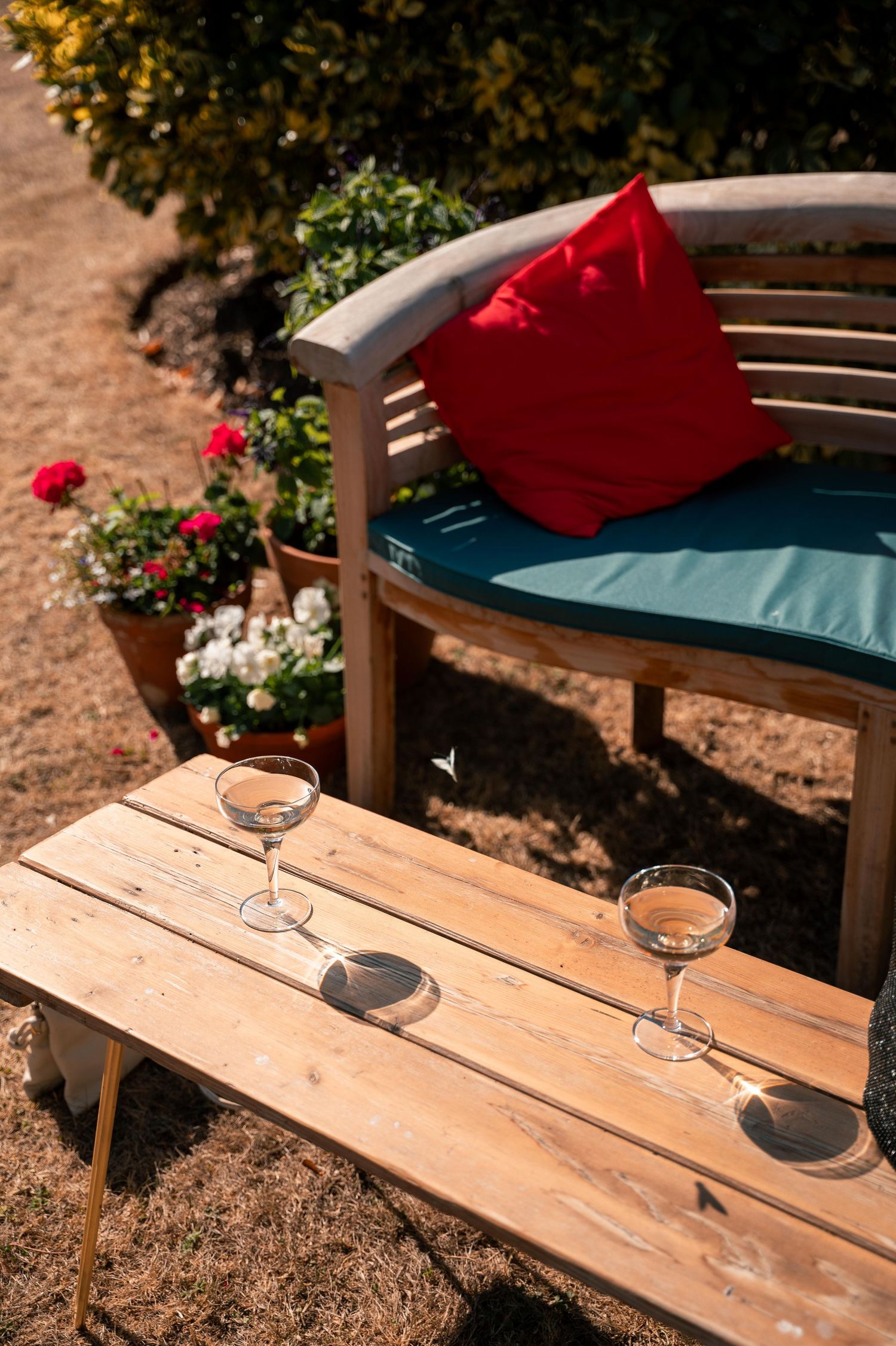 Two wine glasses on wooden table next to curved garden bench with red cushion and blue seat pad in sunny outdoor setting.