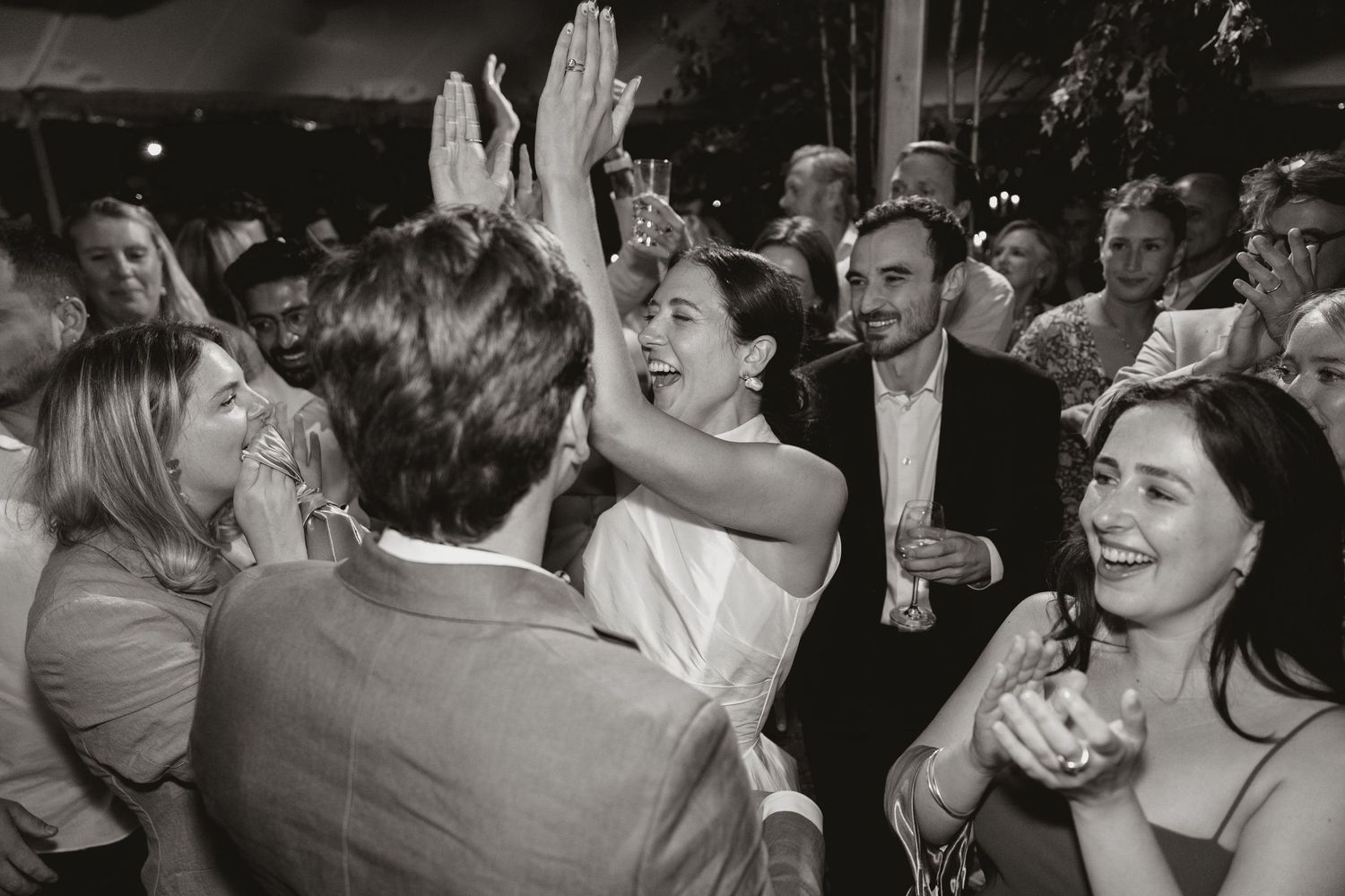 A lively black and white photo of people dancing and celebrating at what appears to be a wedding reception or party.