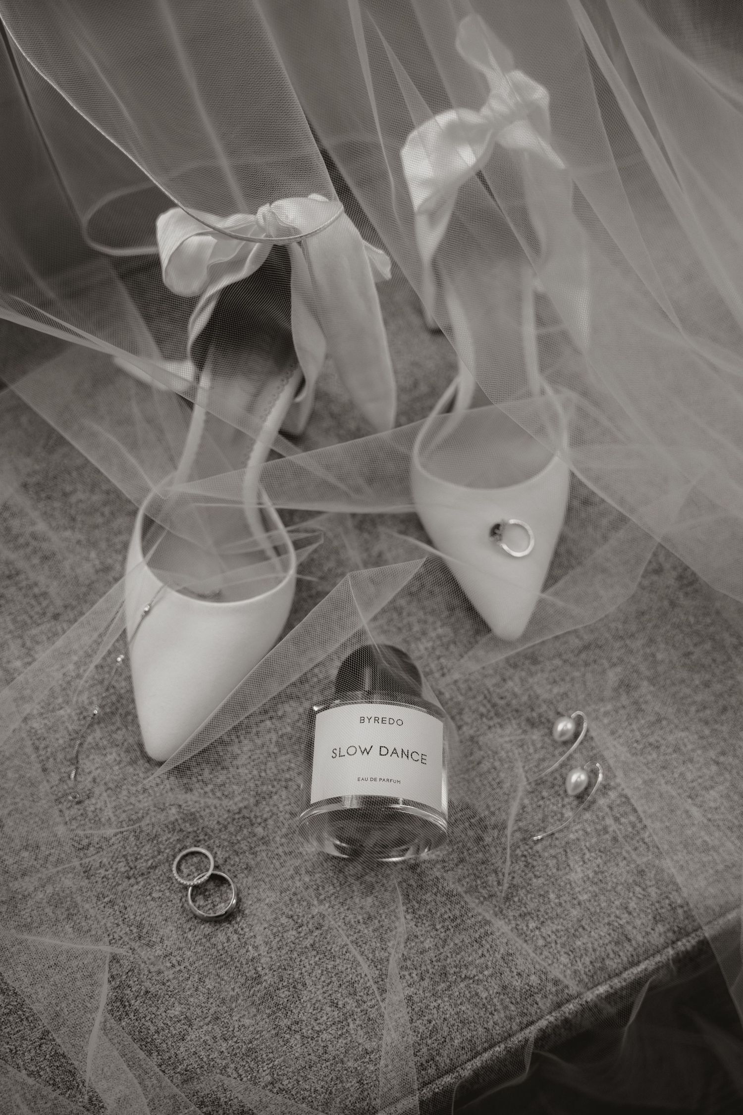 Black and white wedding detail photos showing bridal shoes, veil, and rings arranged on a textured surface.
