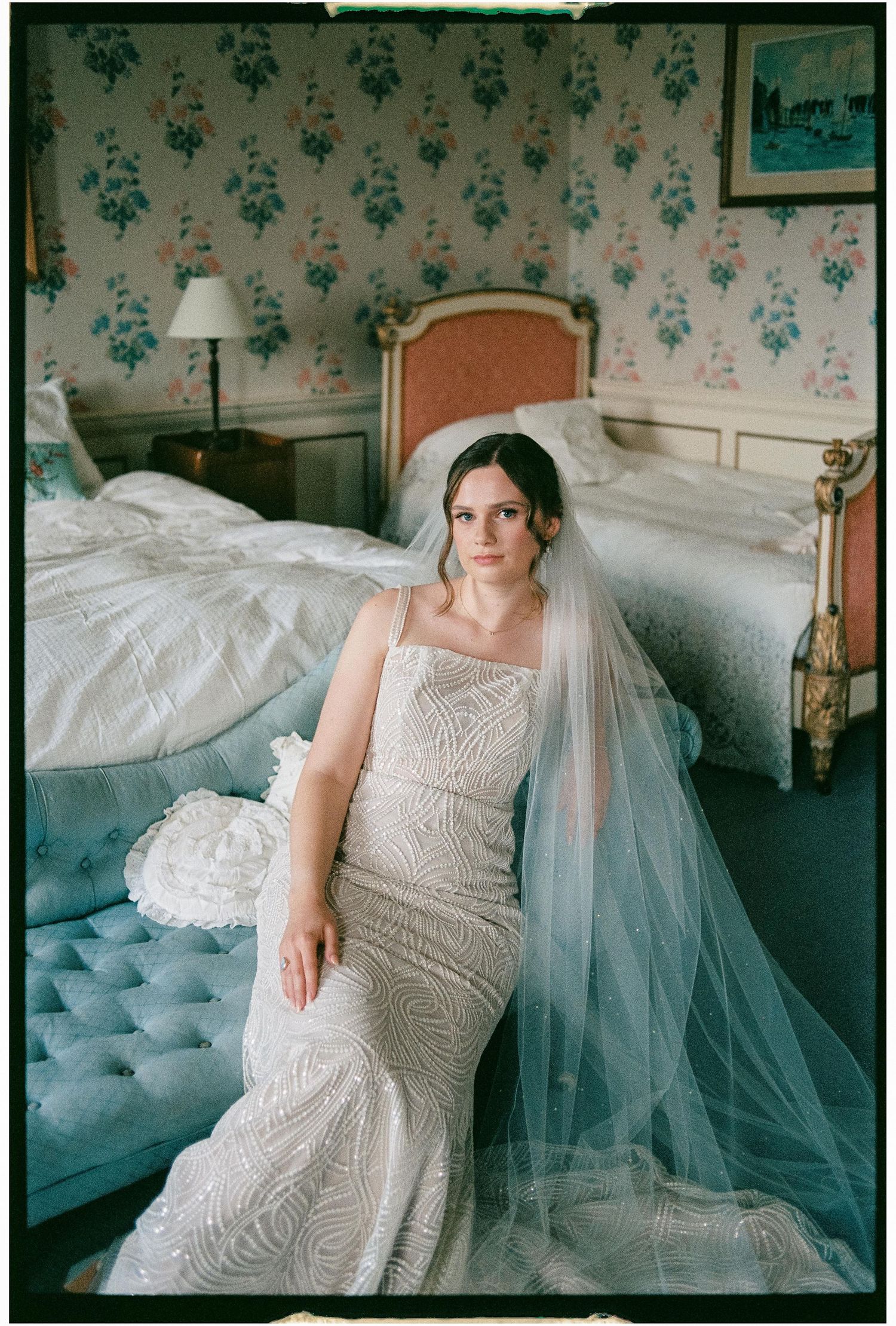 A bride in a fitted lace wedding gown and long veil reclines on a tufted chaise in a room with floral wallpaper.