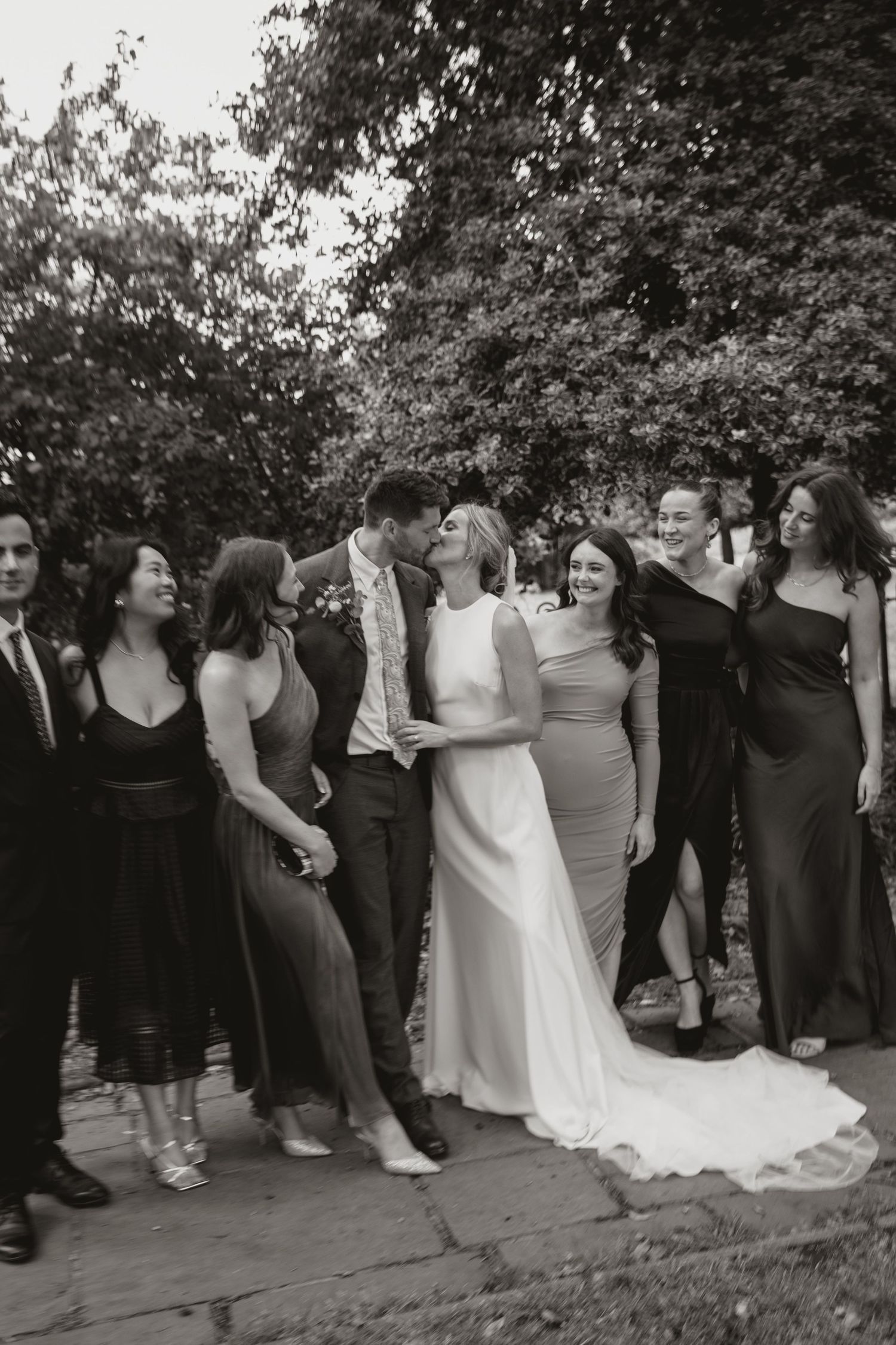 A wedding party stands in a line outdoors under trees during a celebration.