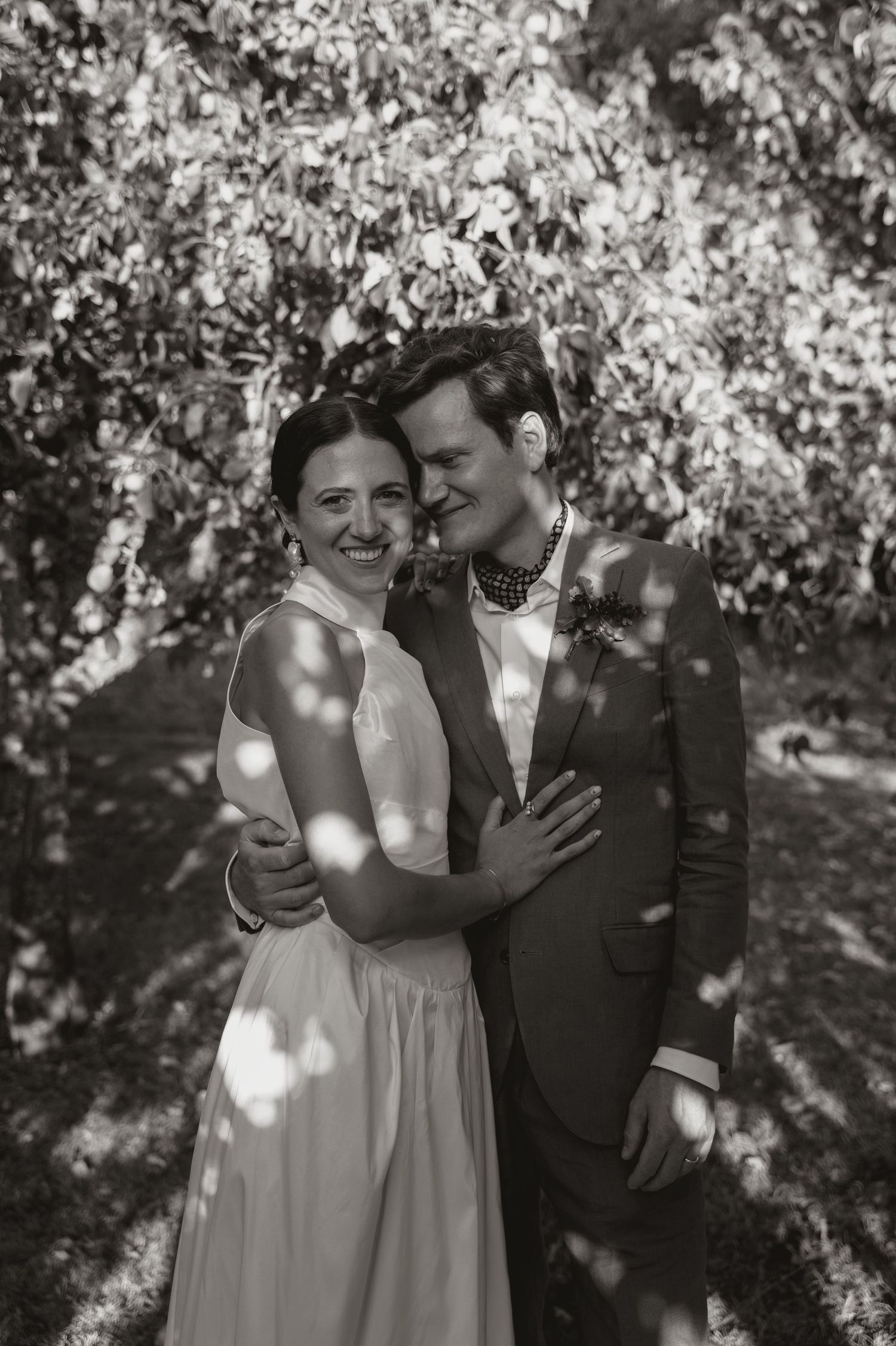 A couple shares intimate moments in a sunlit orchard with blooming trees creating a romantic black and white setting.