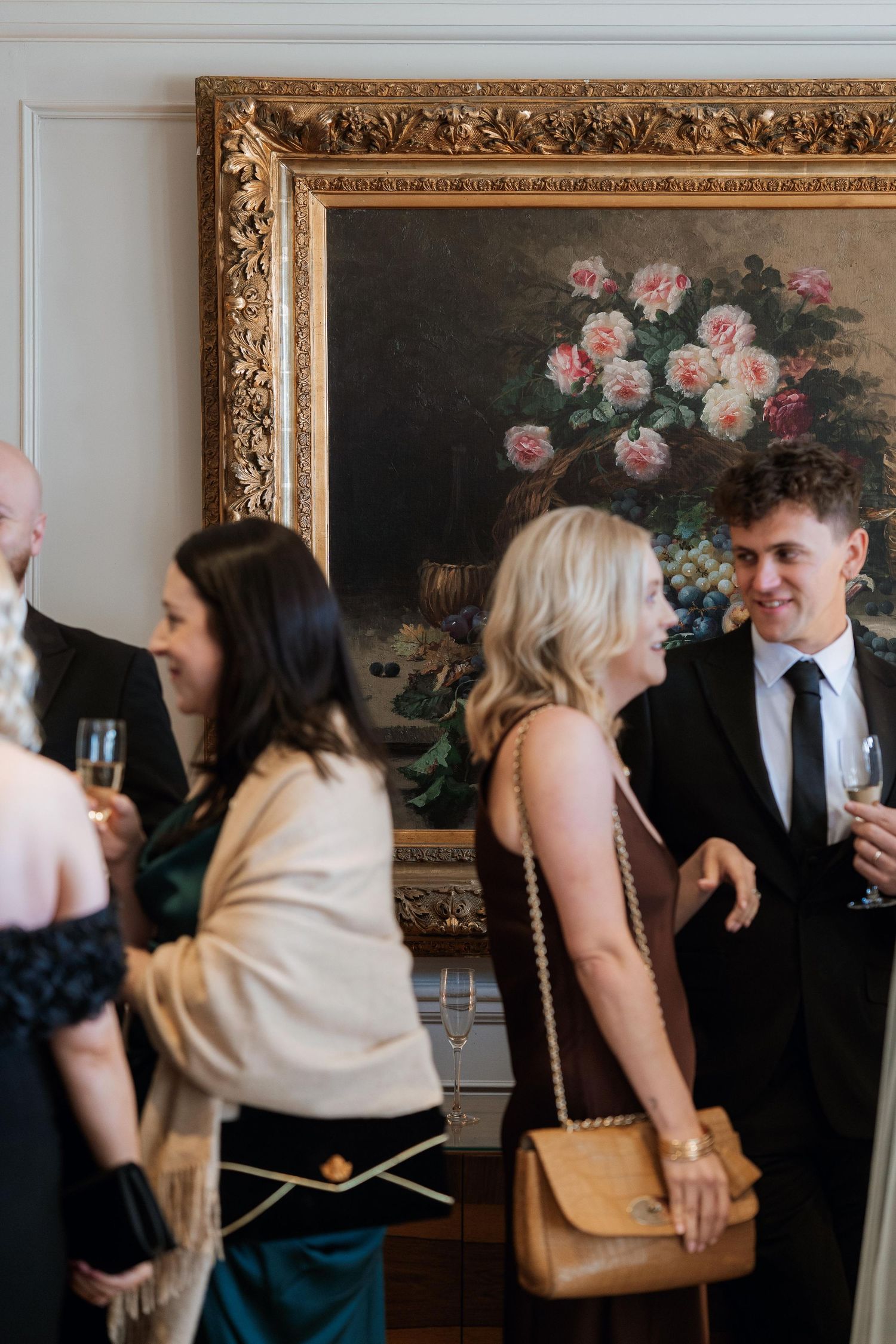 Elegant gathering of guests socializing at an event beneath an ornate gold-framed floral painting.