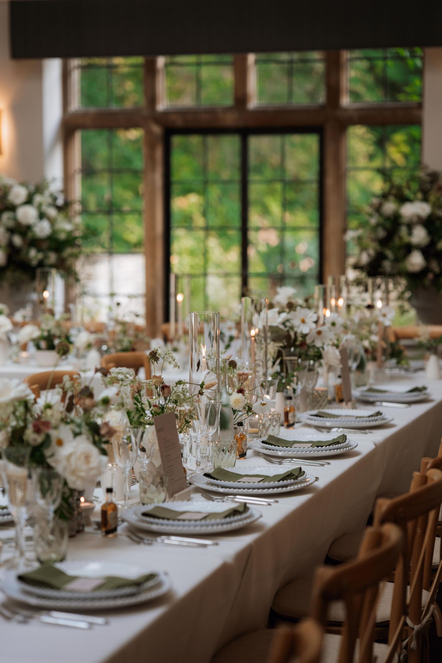 Elegant wedding reception table setting with white flowers, crystal glassware and wooden chairs in natural light.
