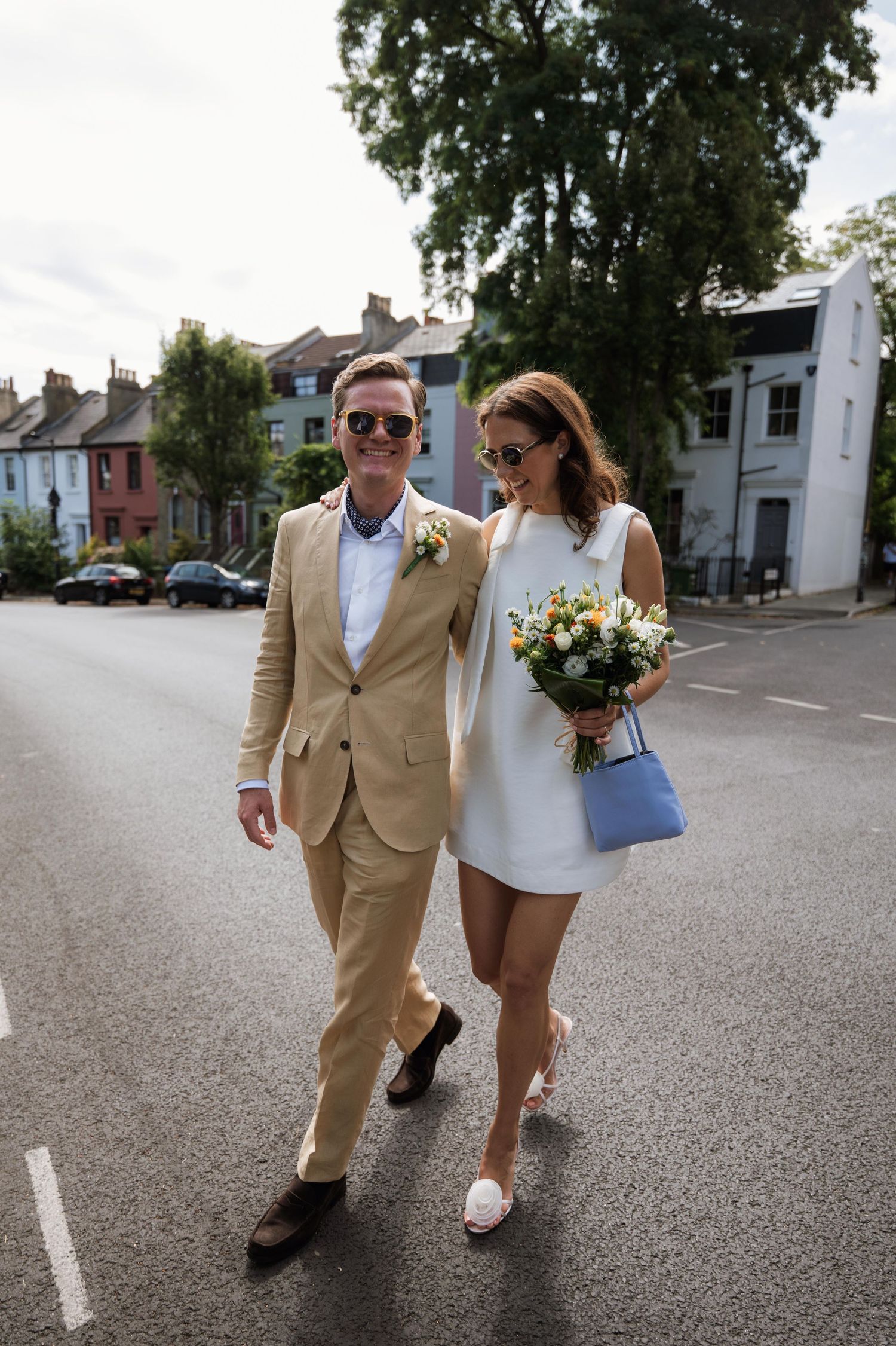 A couple walks down a street, wearing coordinated outfits with a tan suit and white dress, carrying a floral bouquet.