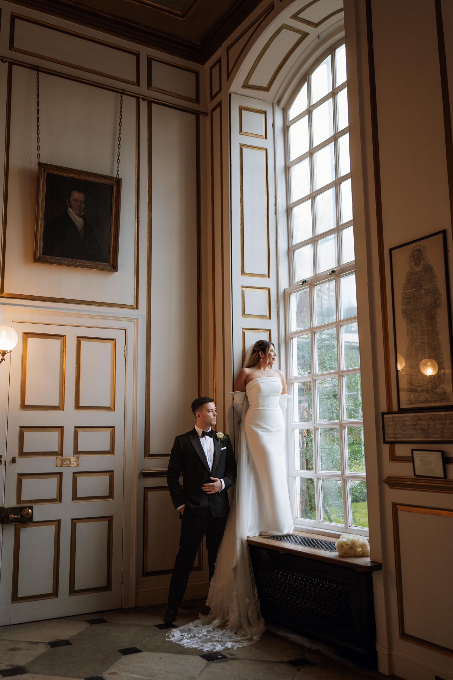 Bride and groom pose in elegant ballroom by tall arched window, groom in tuxedo, bride in white gown with lace train.