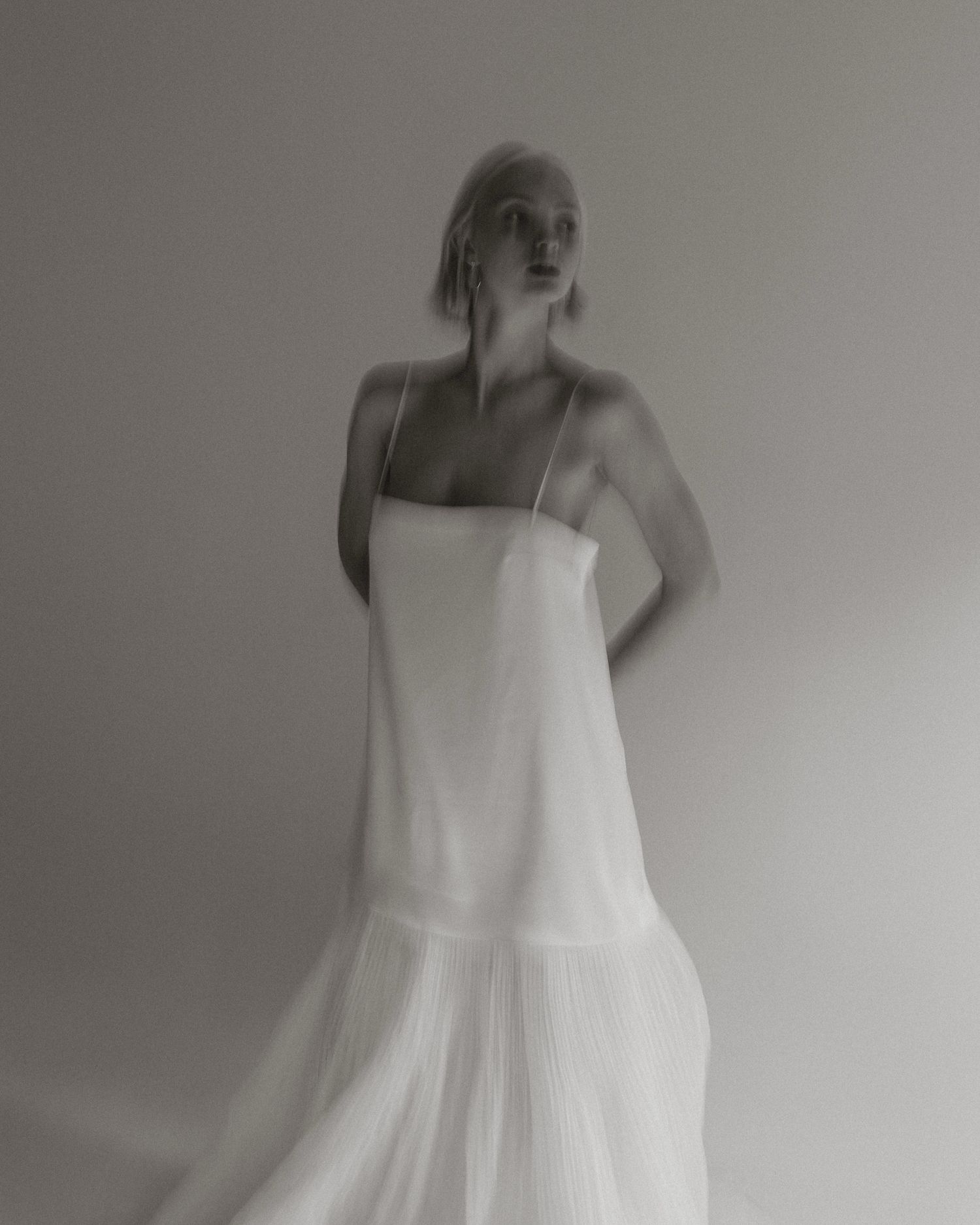 Artistic black and white fashion photograph showing a figure in a flowing white dress against a minimalist background.